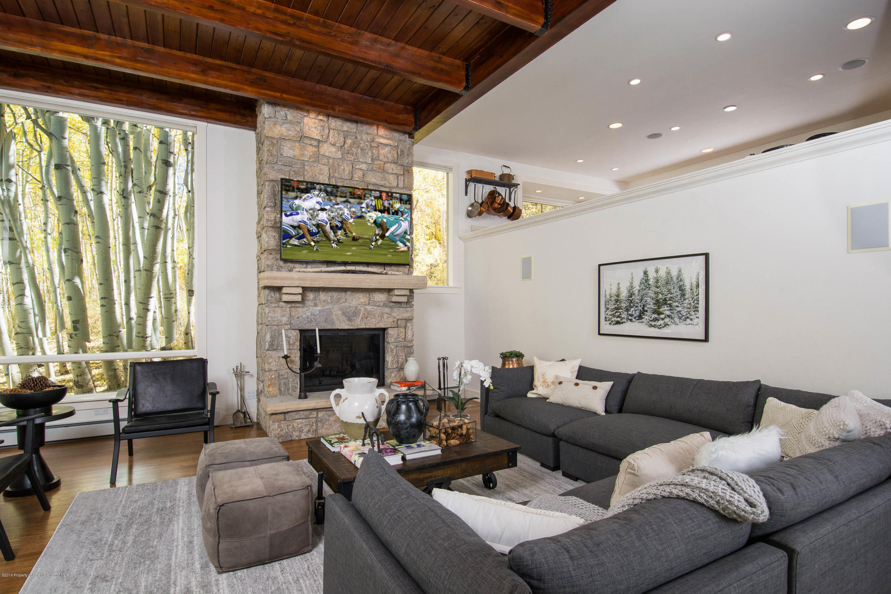 Living room with a stone fireplace, gray sectional, vaulted wood ceiling, and windows overlooking aspens.
