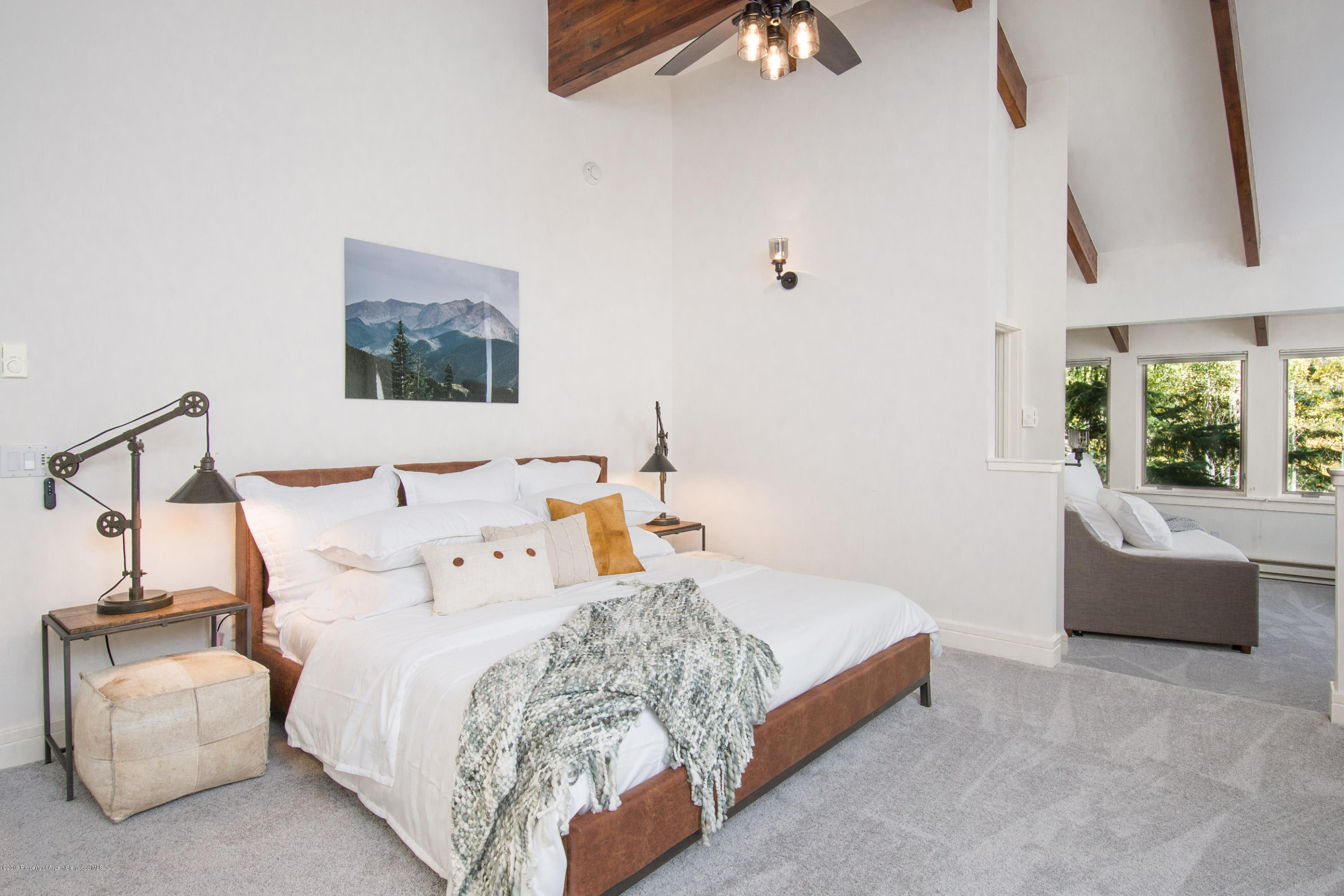 Bedroom with a white bed, exposed wood beams, and a sitting area beside leafy windows.