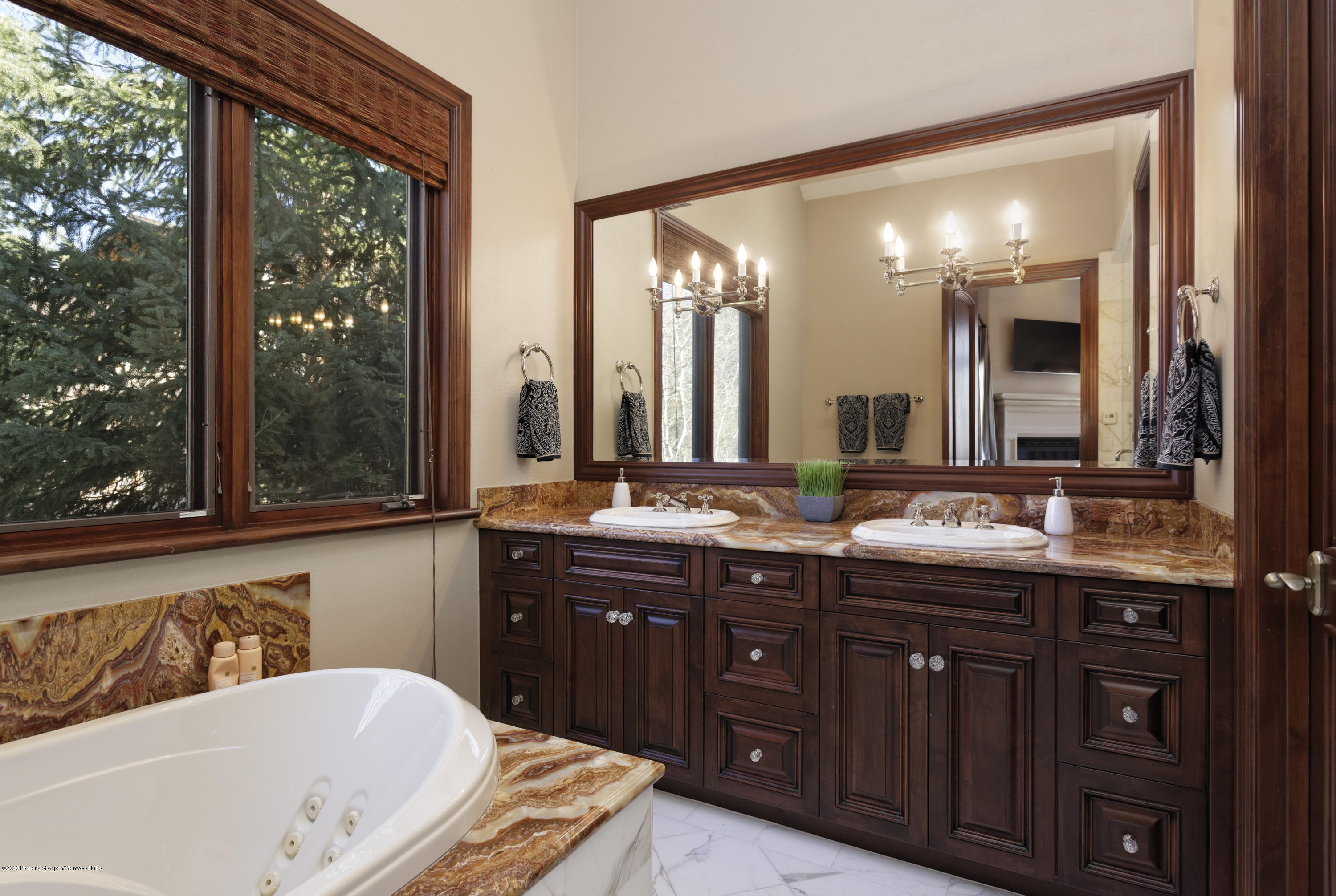 Bathroom with jetted tub, double-sink dark wood vanity, and window overlooking evergreen trees.