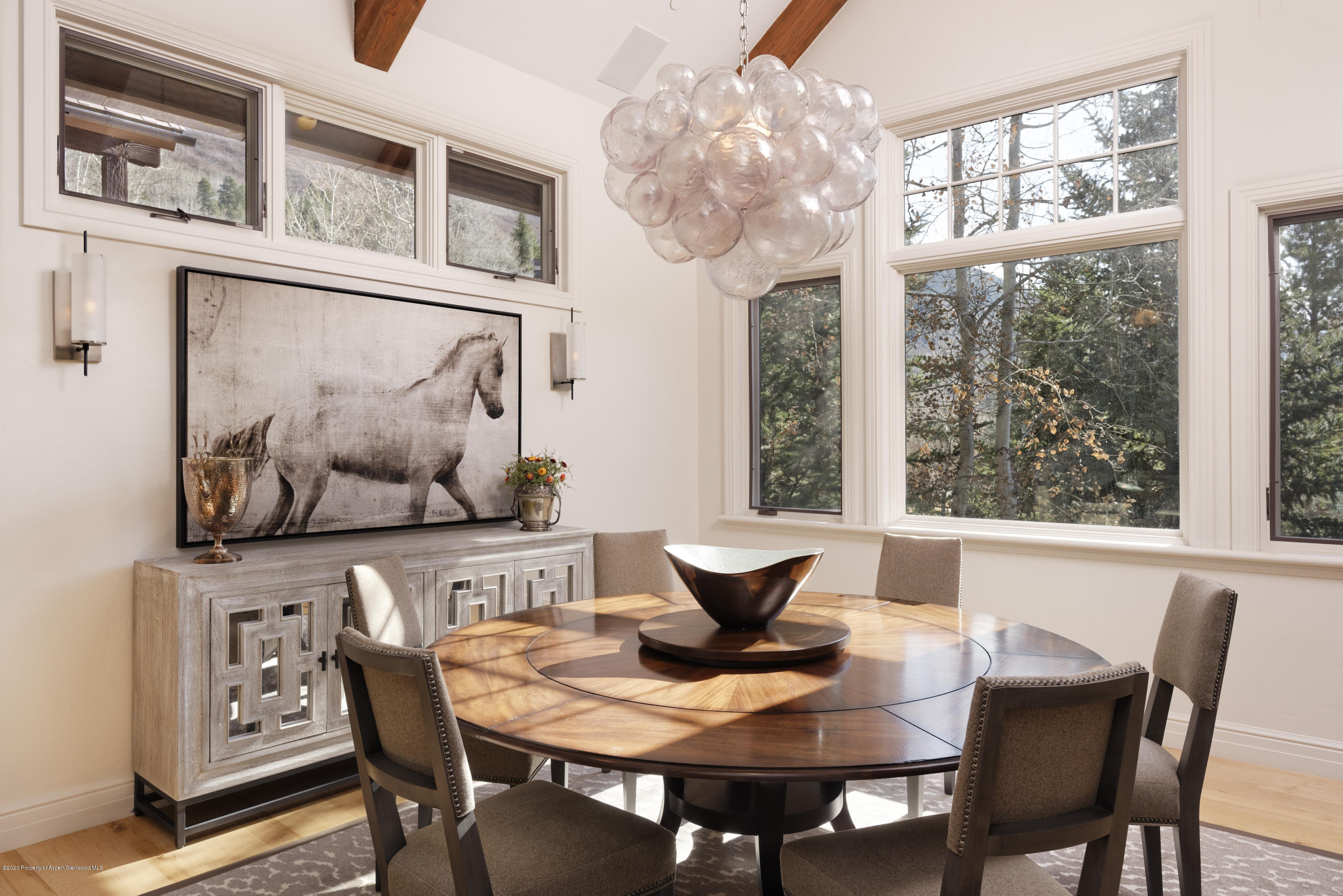 Dining room with a round wood table, glass chandelier, horse artwork, and large windows overlooking trees.
