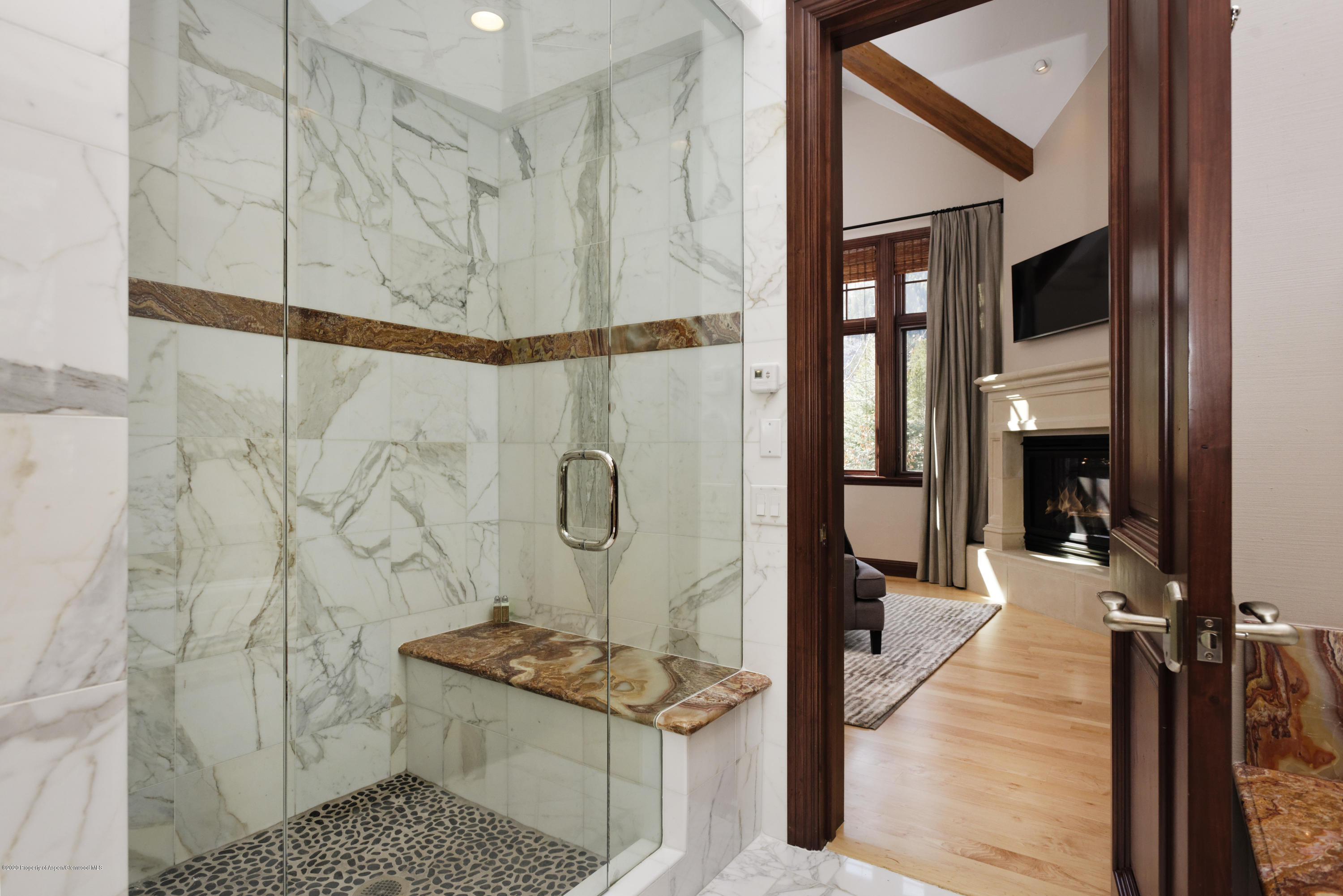 Marble walk-in shower with a stone bench, opening onto a room with a fireplace.