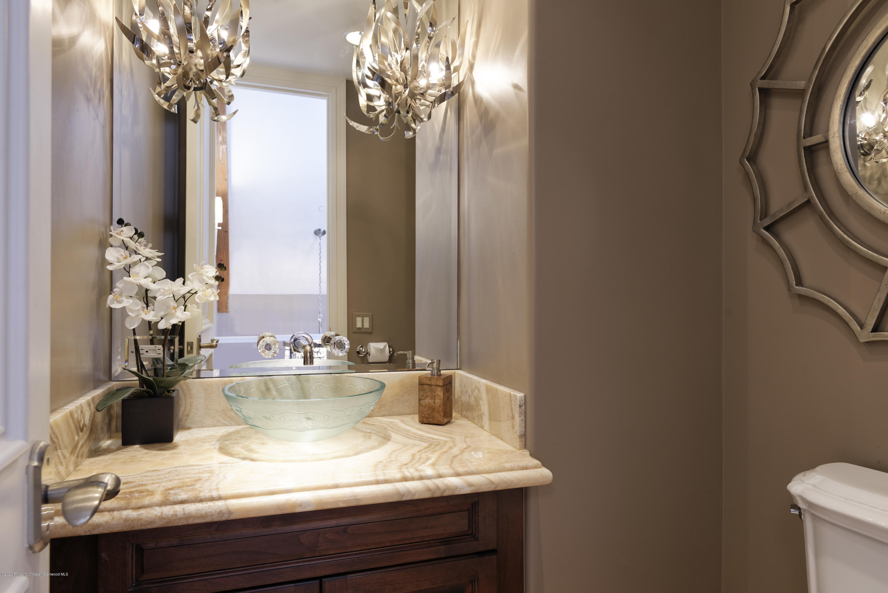 Bathroom vanity with a glass vessel sink, pale stone counter, orchids, and sculptural pendant lights.