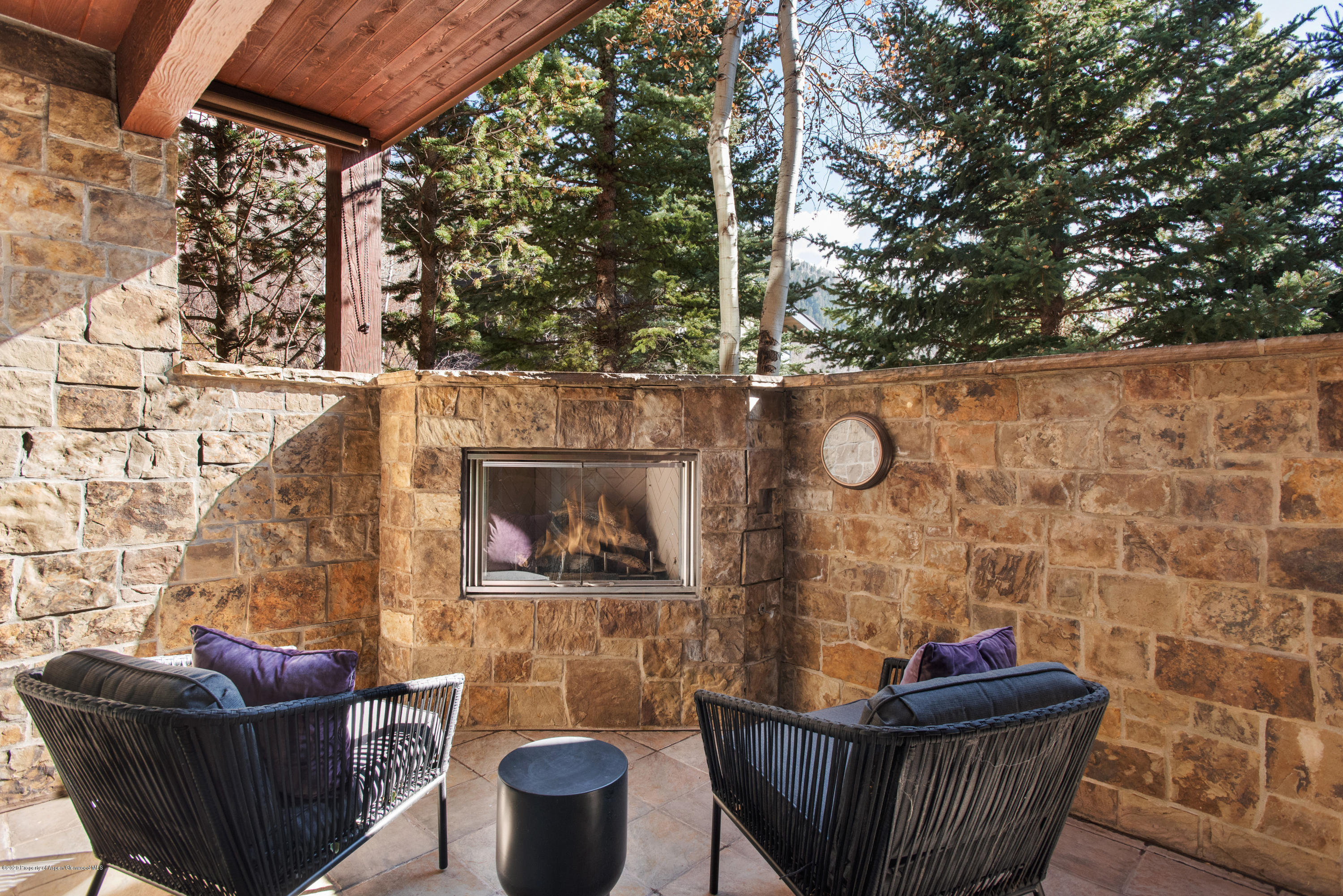 Covered stone patio with a fireplace, woven lounge chairs, and trees beyond the walls.