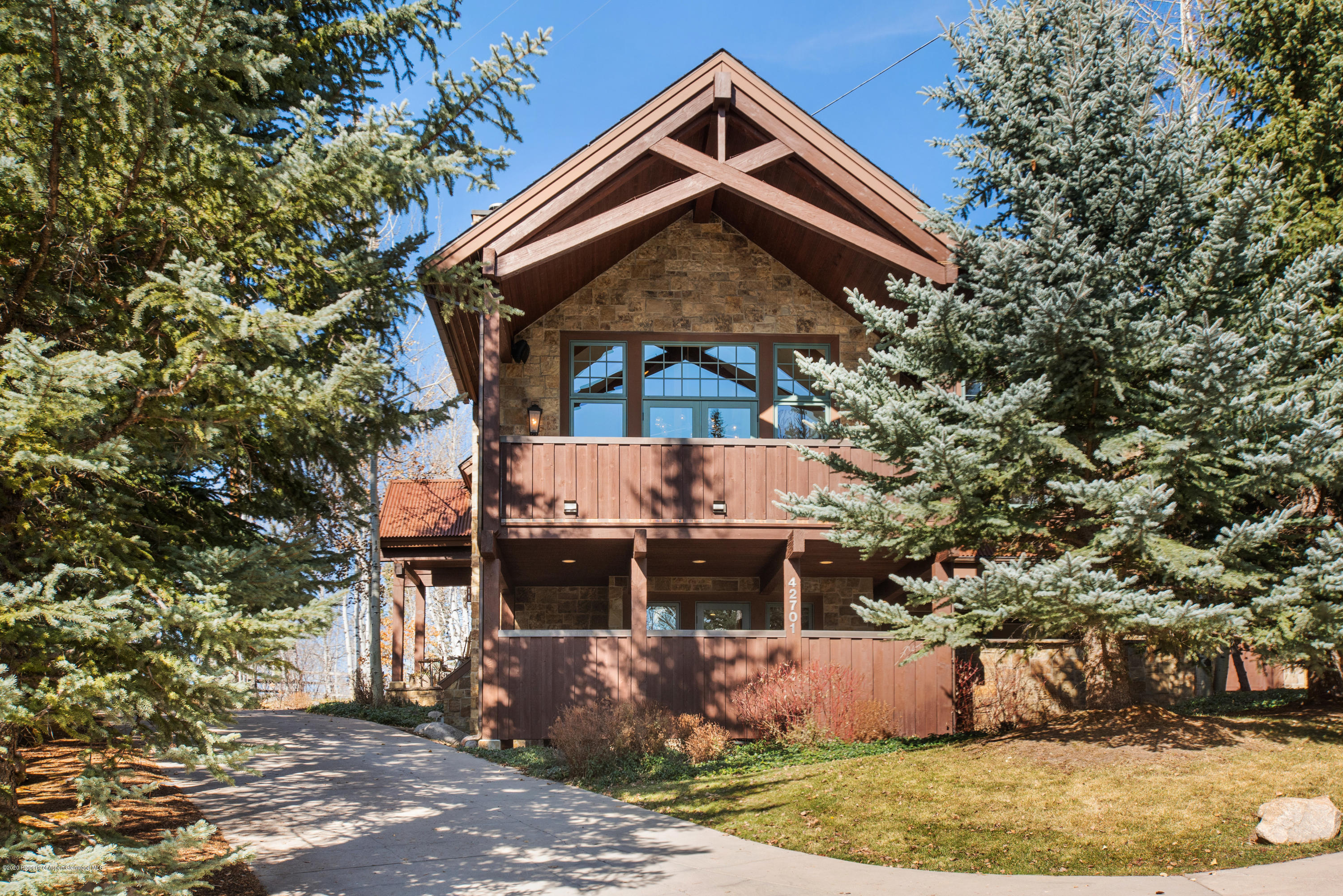 Mountain-style home with a stone facade and timber gable, framed by evergreen trees beside the driveway.