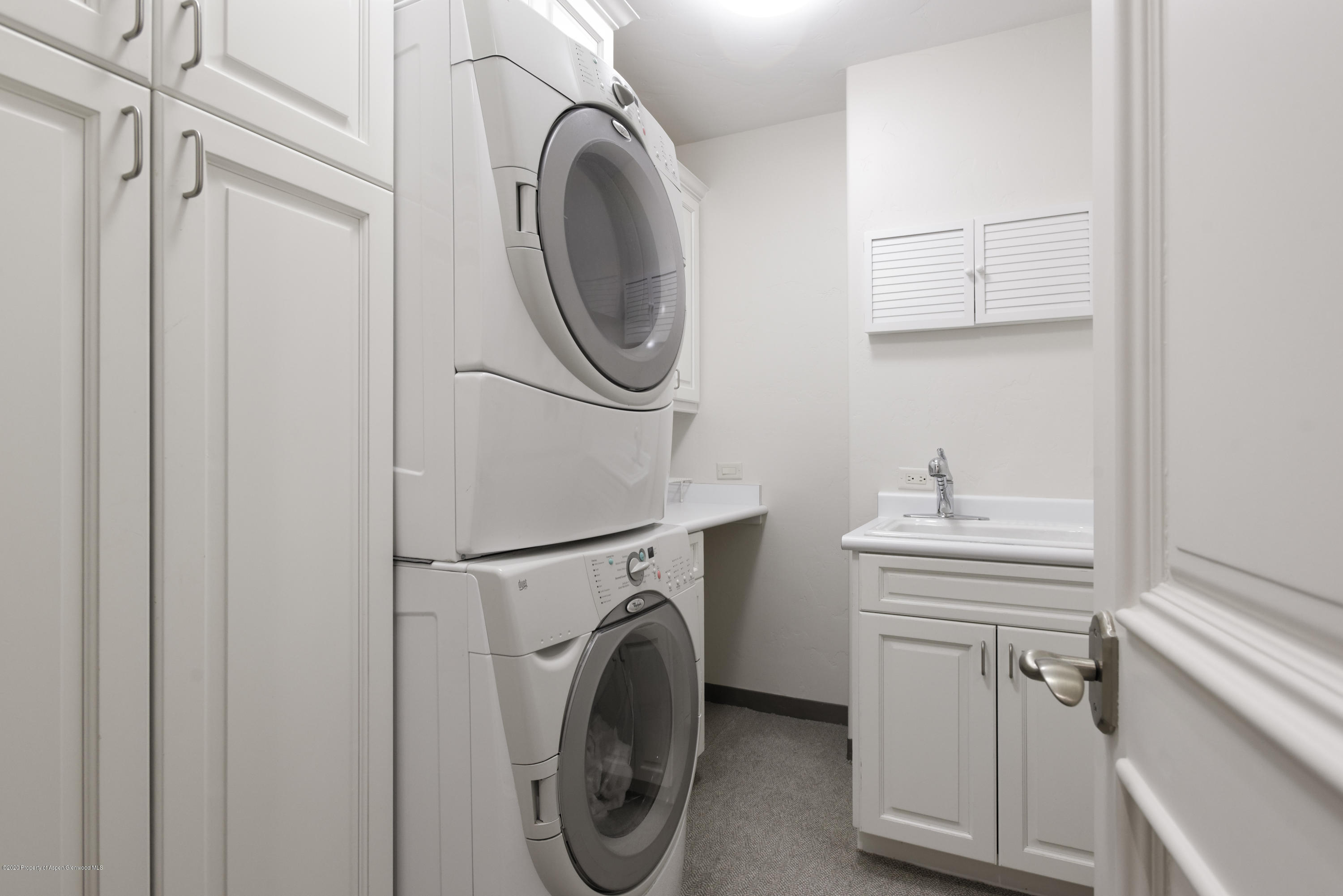 Laundry room with stacked front-loading washer and dryer beside white cabinets and a utility sink.