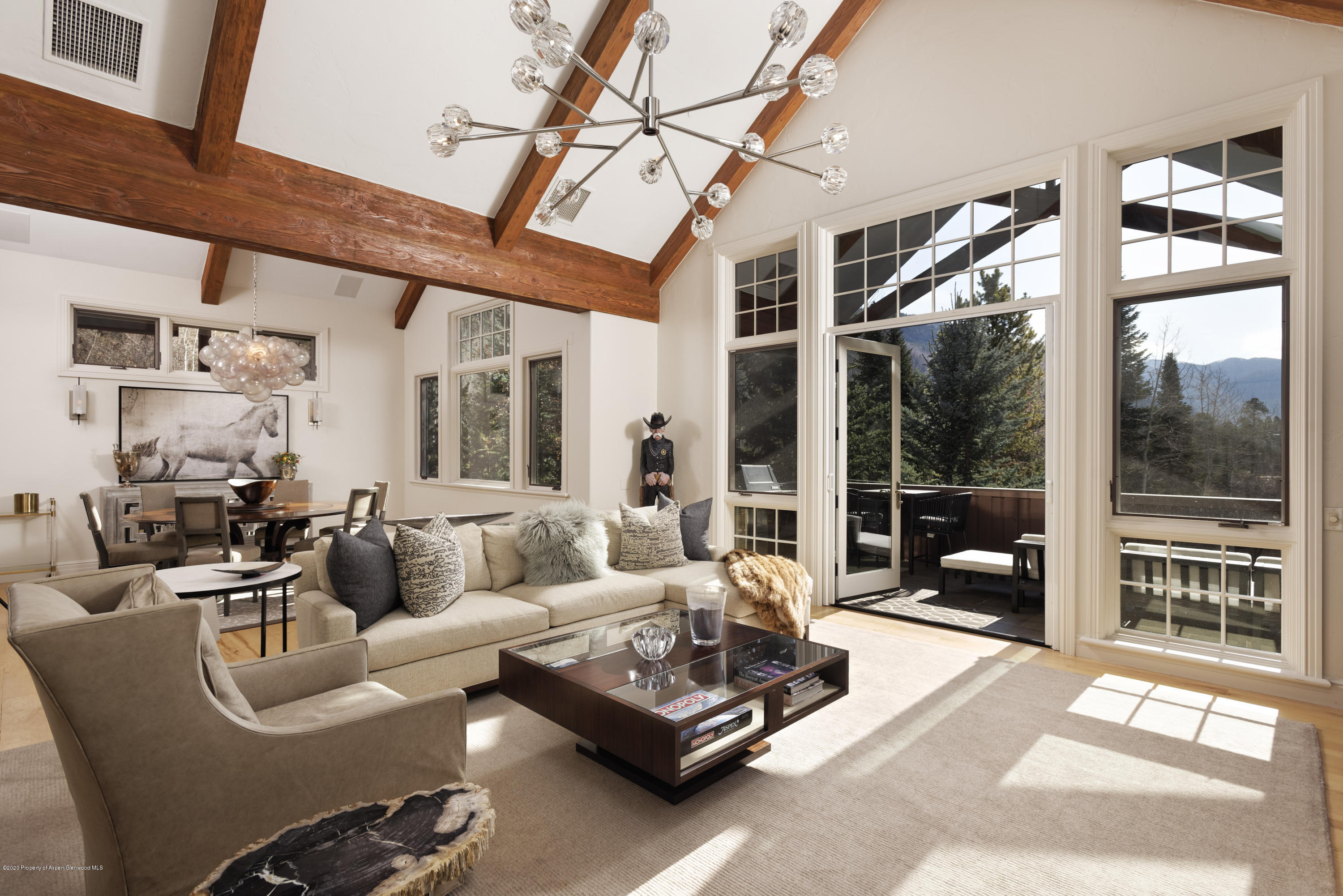 Furnished living and dining room with exposed timber beams, tall windows, and a balcony overlooking mountains.