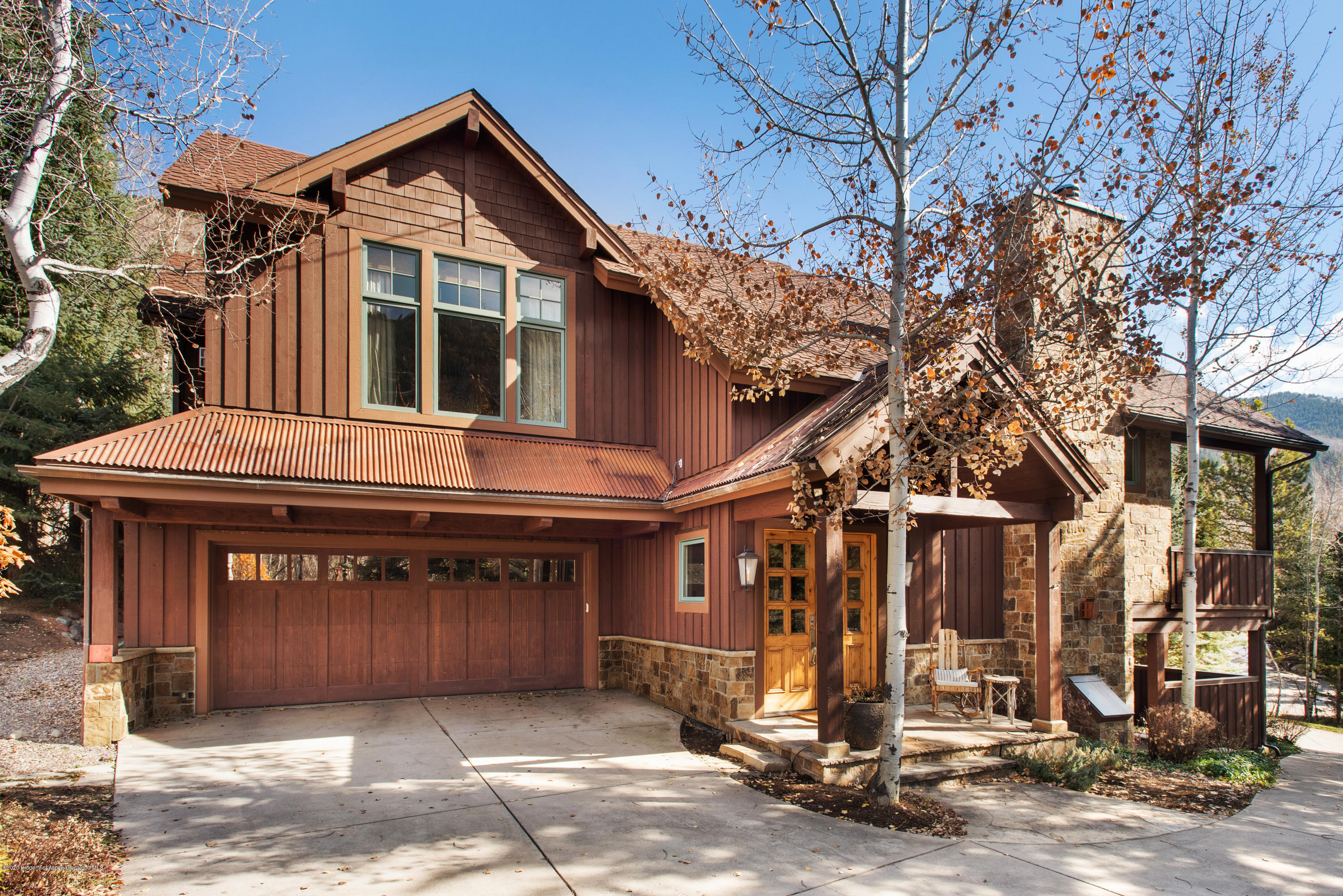 Two-story brown home with a large garage, stone accents, and a covered entry among autumn birch trees.