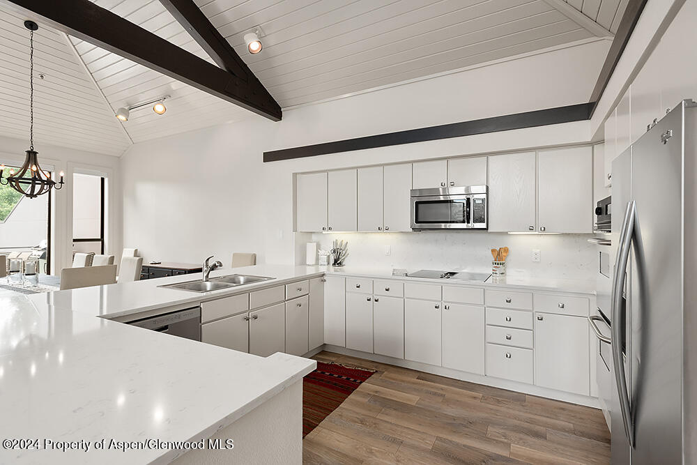Modern white kitchen with a large island, stainless steel appliances, and vaulted ceiling with dark beams.