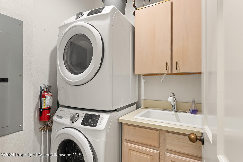 Laundry room with stacked washer and dryer beside a utility sink and light wood cabinets.