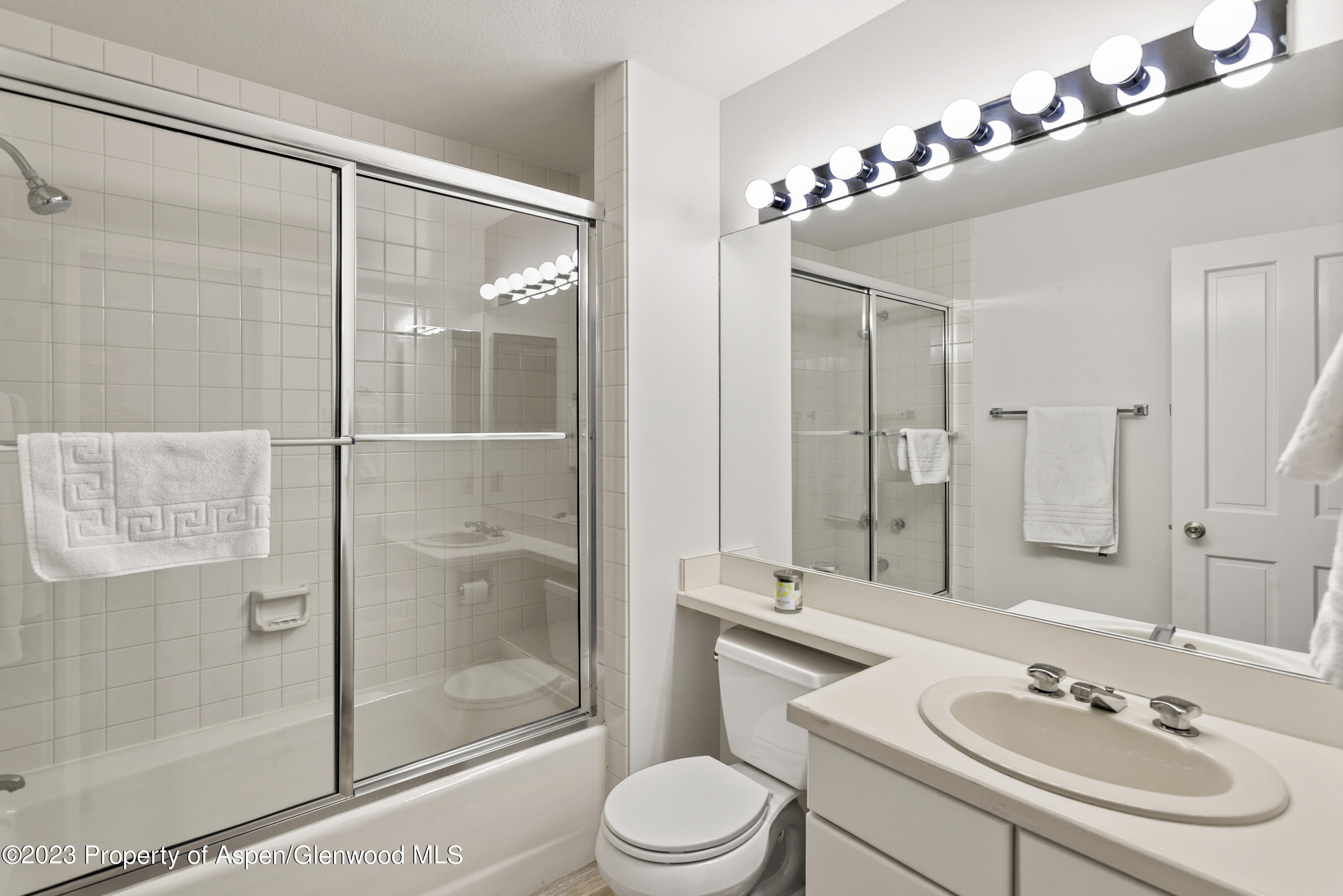 Bathroom with glass-enclosed tub and shower, large mirror, white vanity, and toilet.
