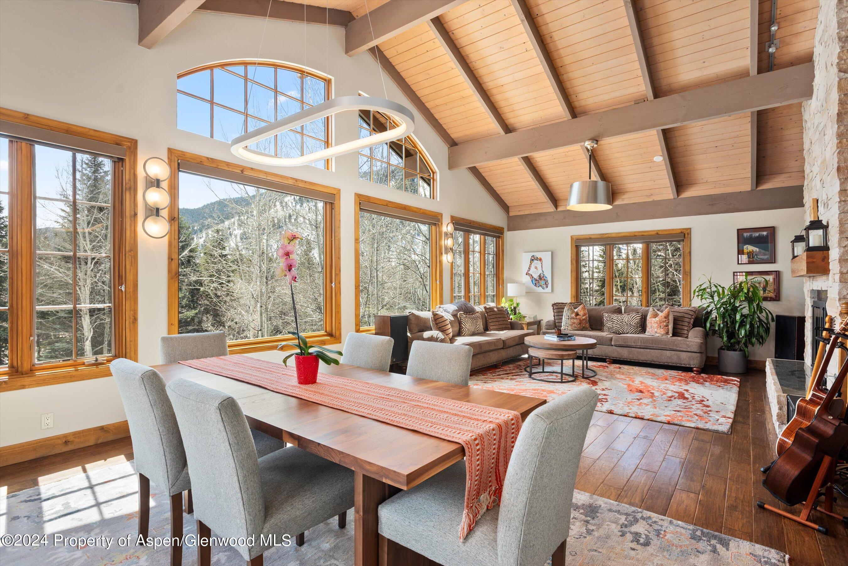 Open-plan living and dining room with vaulted wood ceiling, large windows, and mountain views.