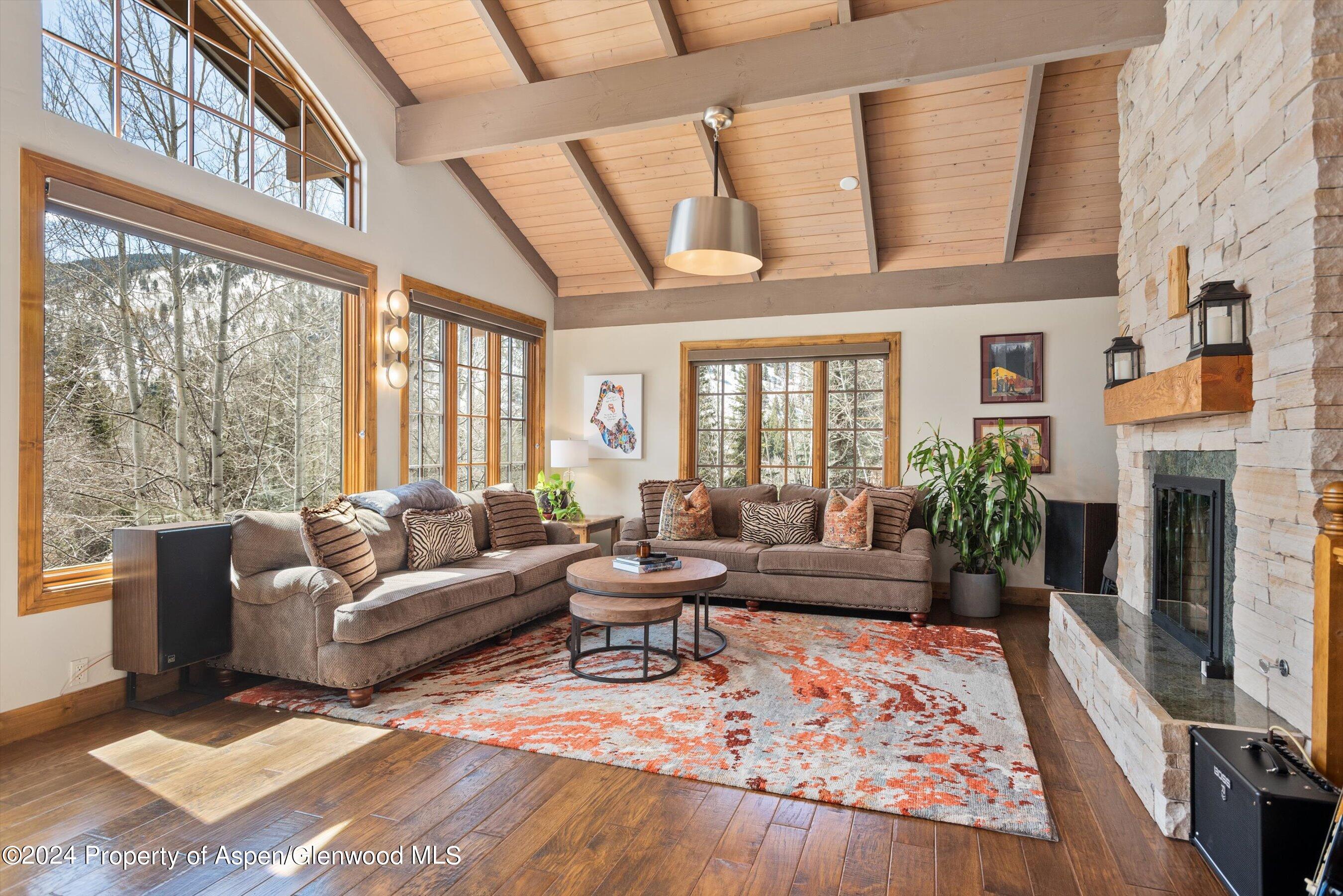 Living room with a vaulted wood ceiling, large windows, and a tall stone fireplace.