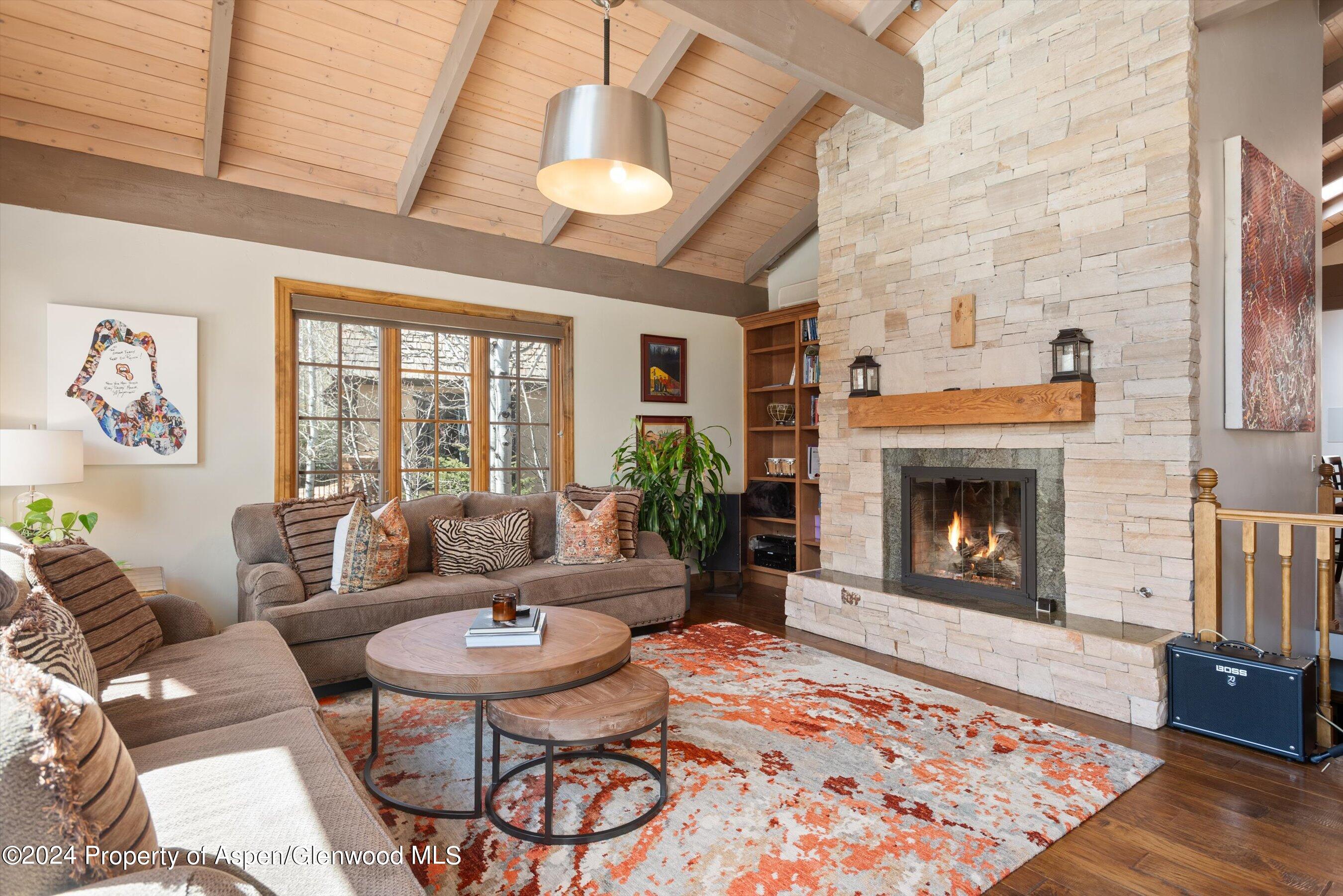 Living room with a vaulted wood ceiling, stone fireplace, broad windows, and orange-patterned rug.
