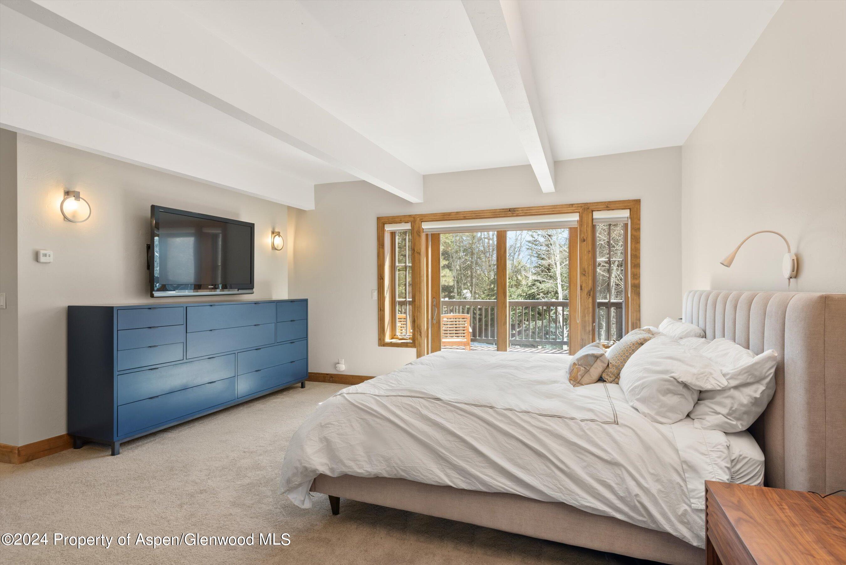 Bedroom with a large bed, blue dresser, and wood-framed windows overlooking a tree-lined deck.