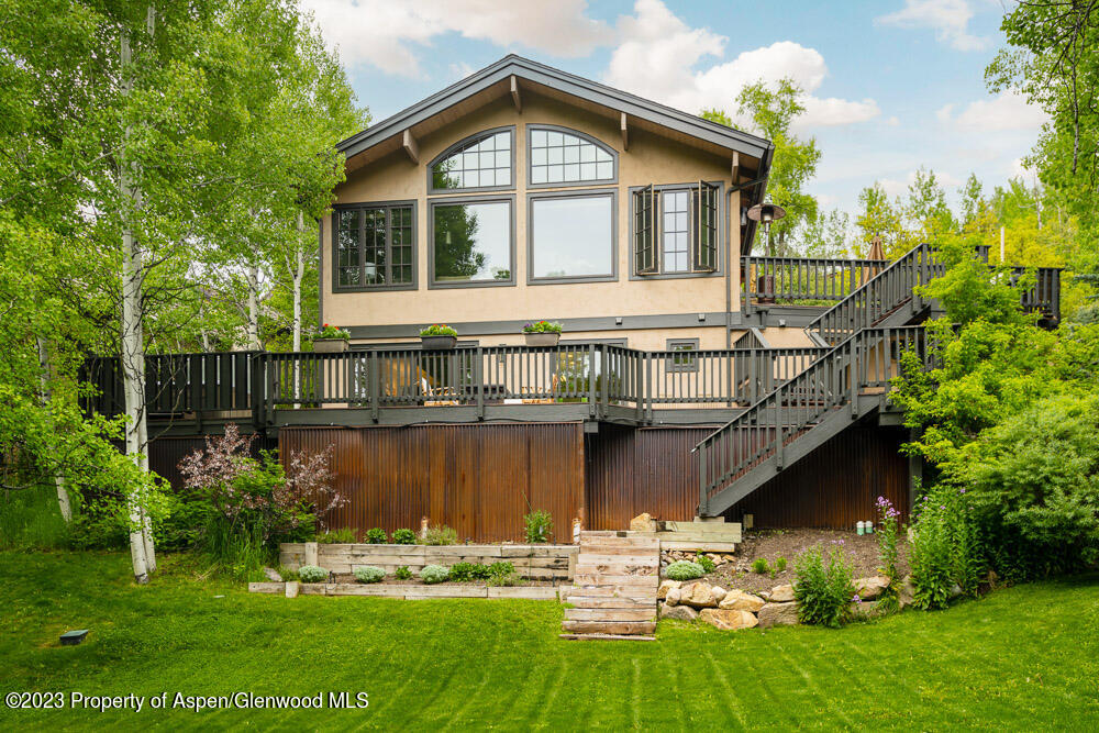 Home with an elevated deck and exterior stairs, surrounded by mature trees and landscaped garden beds.