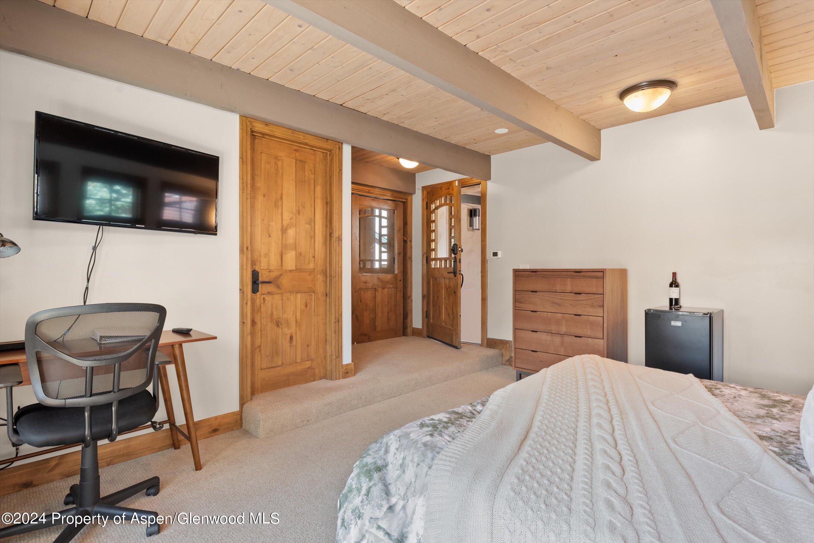 Furnished bedroom with exposed wood ceiling beams, a wall-mounted television, and a desk and chair.