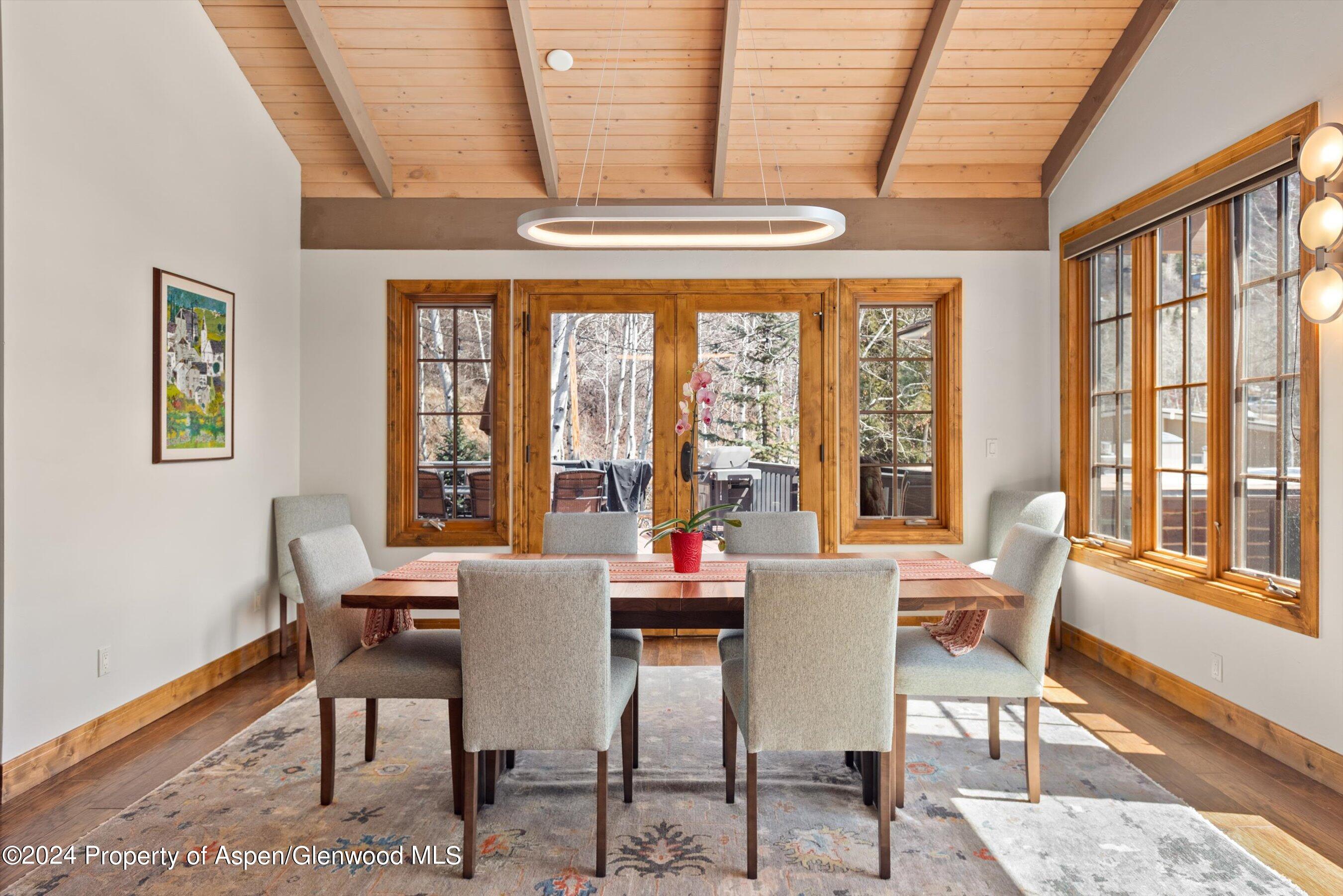 Dining room with a long wooden table, vaulted wood ceiling, and large windows opening toward a deck.