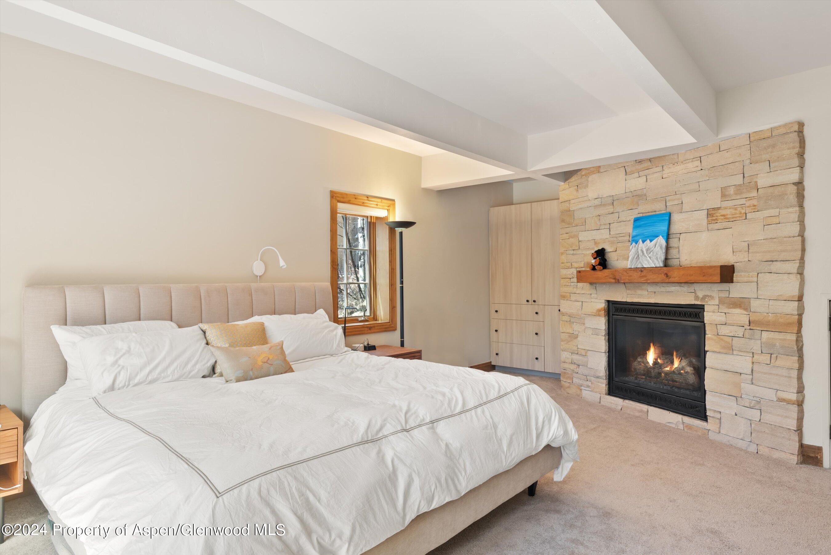 Furnished bedroom with a large white bed beside a stone fireplace with a lit fire.