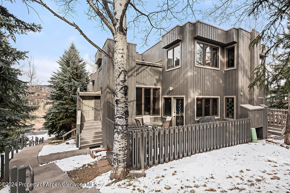 Two-story gray-sided home with a fenced patio and snowy yard among evergreen trees.