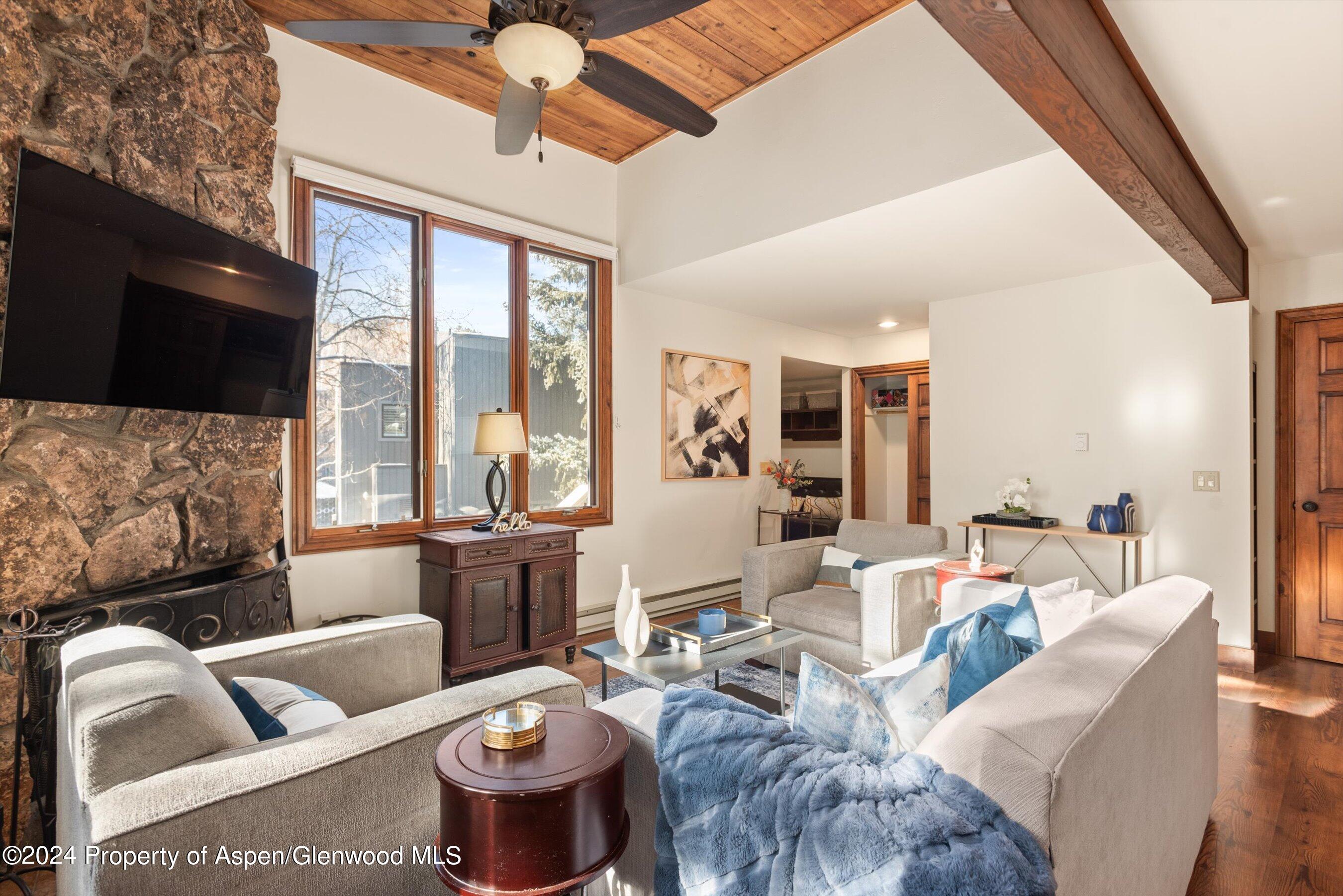 Living room with a stone fireplace, tall windows, wood-plank ceiling, and gray sofas with blue accents.