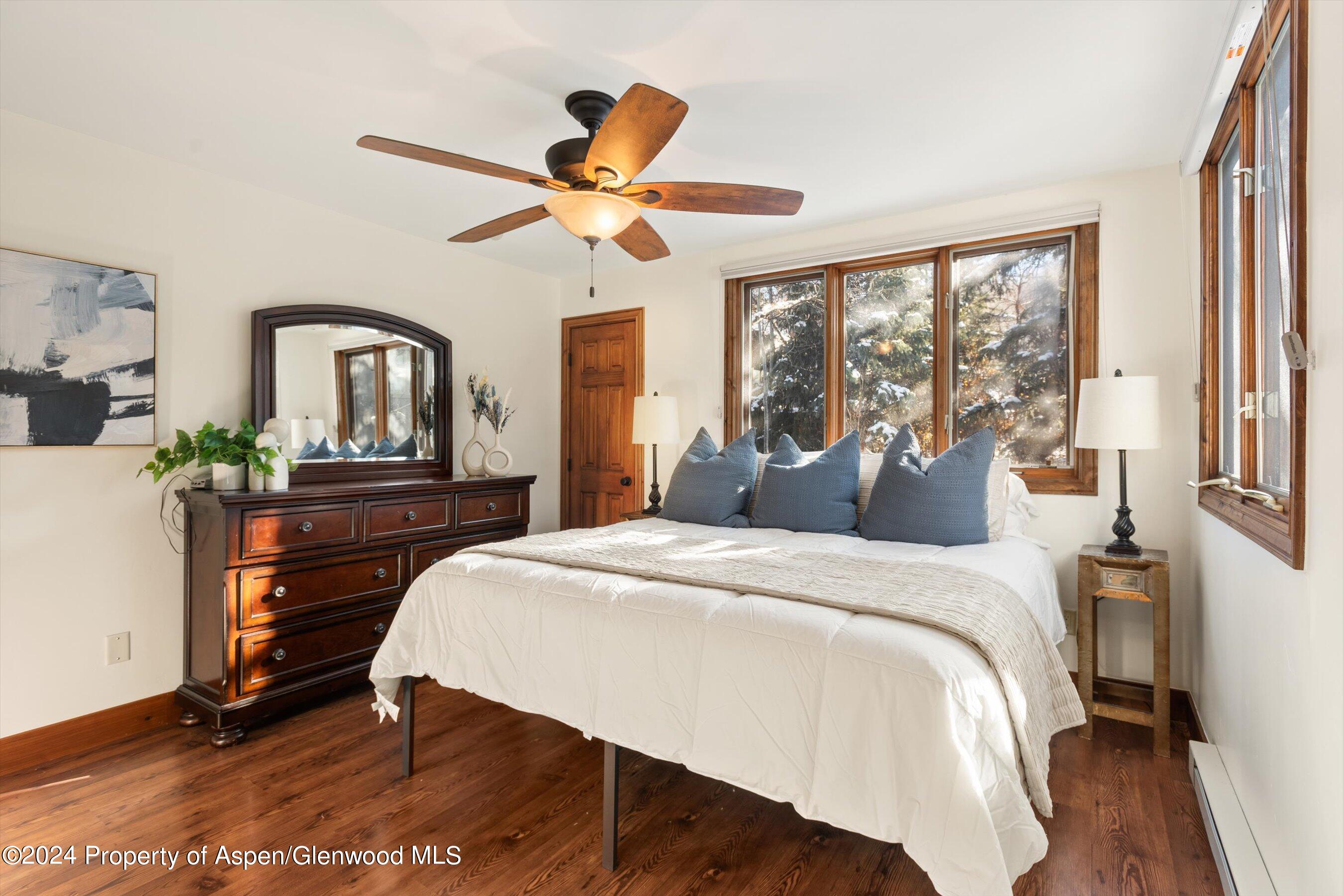 Furnished bedroom with a white bed, blue pillows, dark wood floors, and windows overlooking snowy trees.