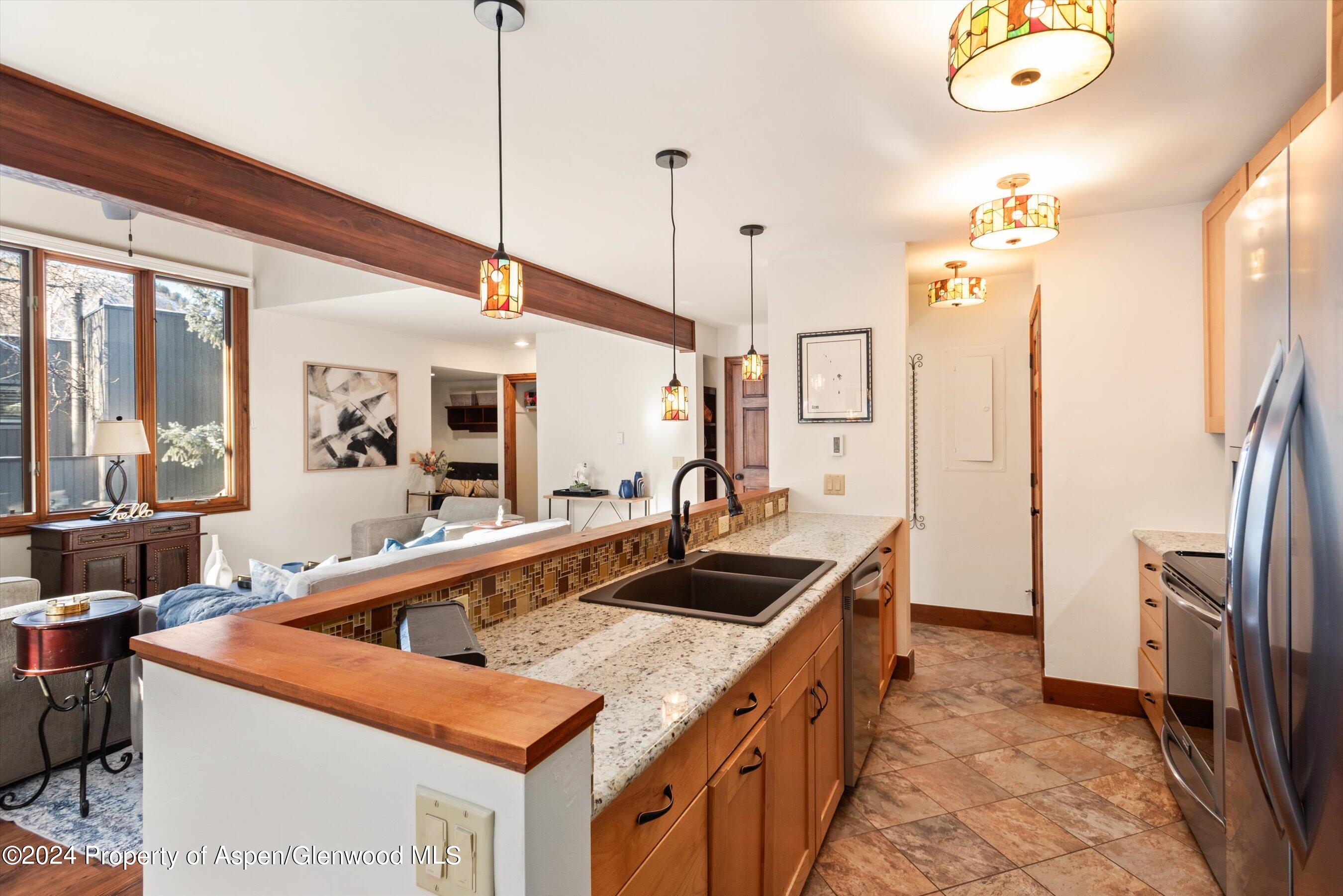 Kitchen with granite counters, wood cabinetry, a dark double sink, and colorful pendant lights opening to a living area.