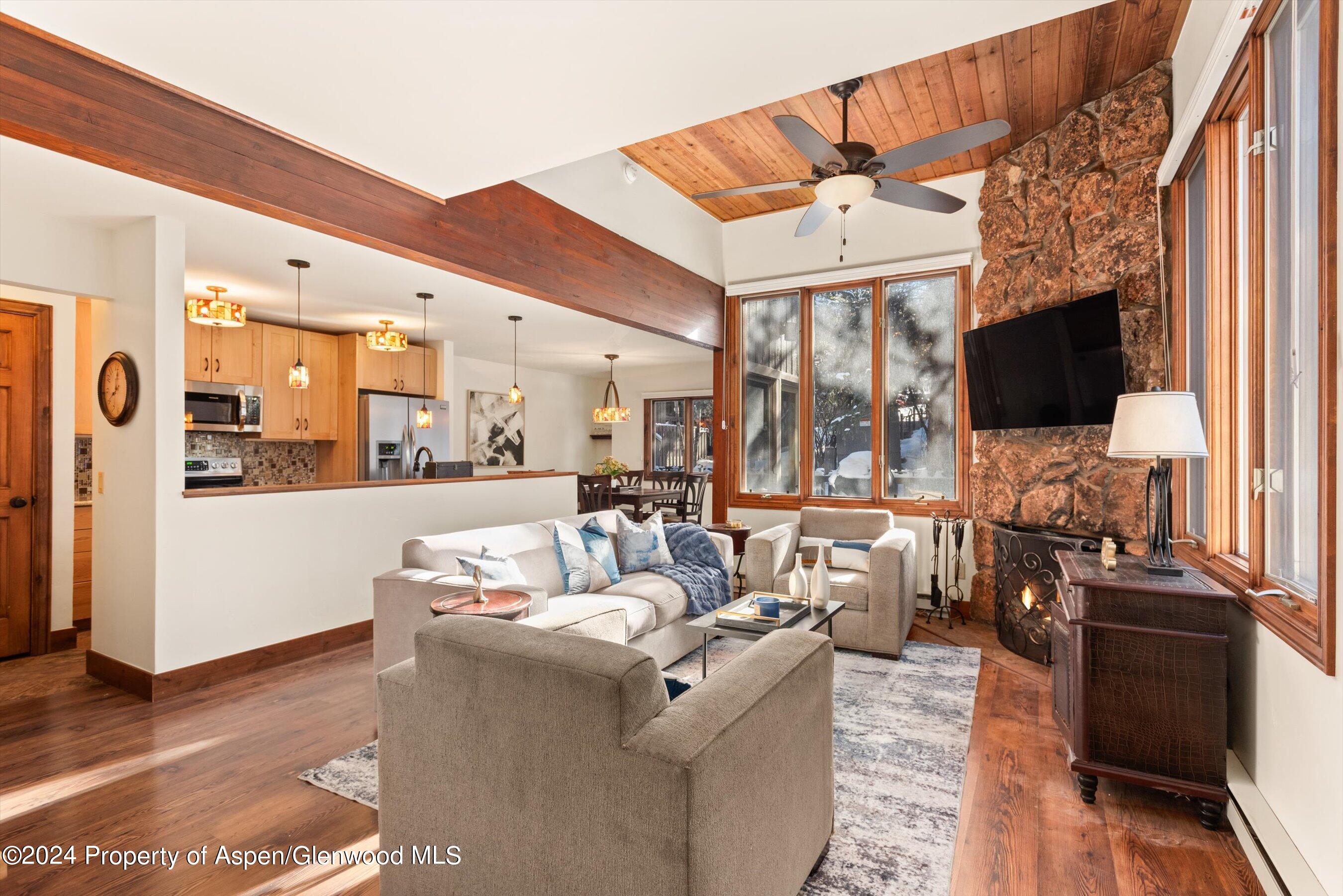 Open-plan living room with a stone fireplace, vaulted wood ceiling, and snowy views through large windows.