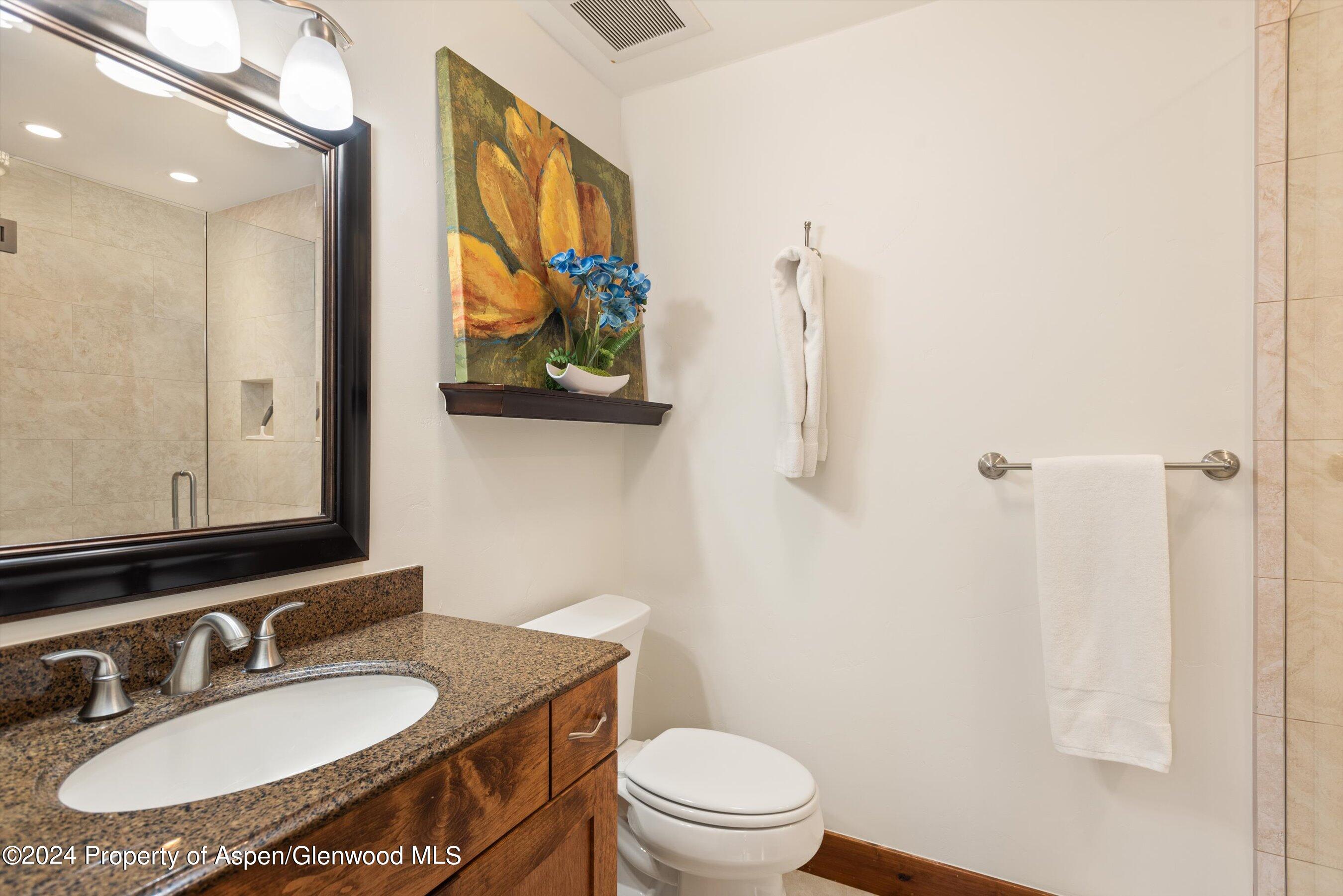 Bathroom with a granite vanity, dark-framed mirror, tiled shower, and colorful floral painting.
