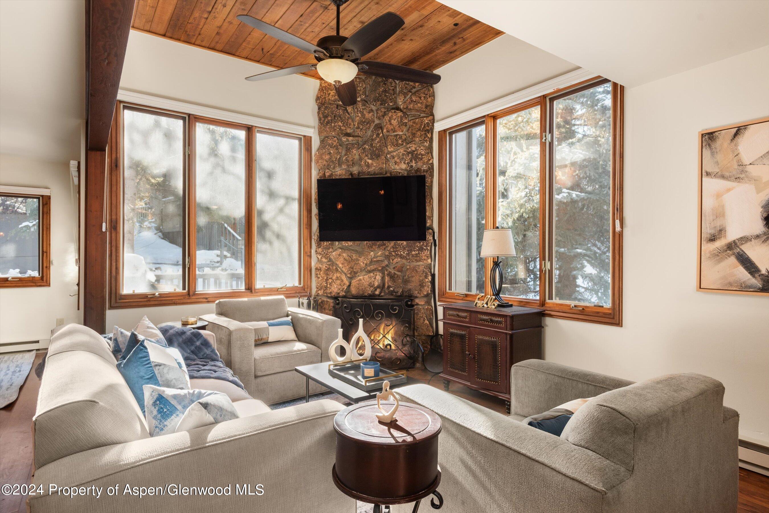 Living room with a tall stone fireplace, wood ceiling, and large windows overlooking snowy trees.