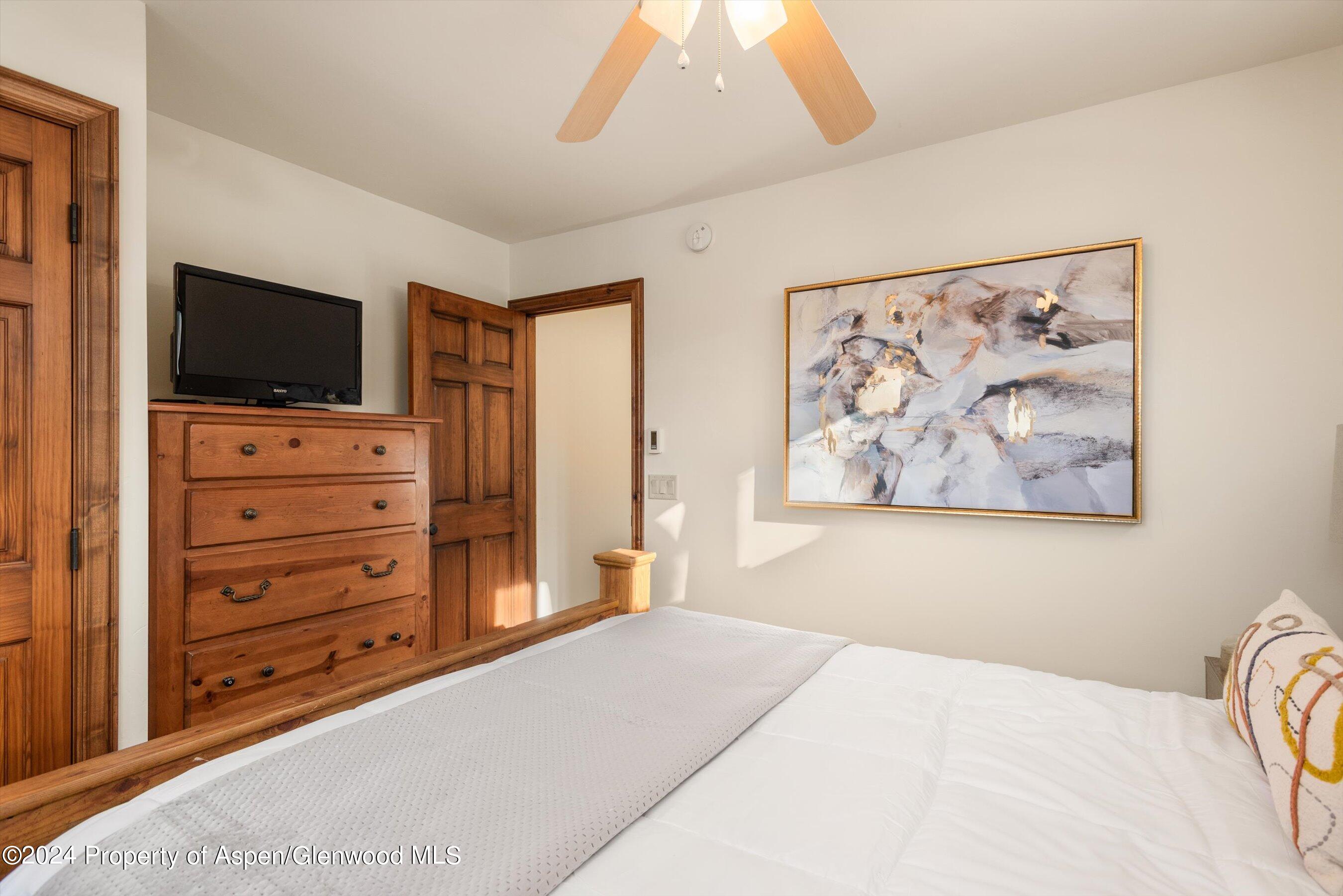 Bedroom with a white bed, rustic wooden furnishings, ceiling fan, and large abstract painting.