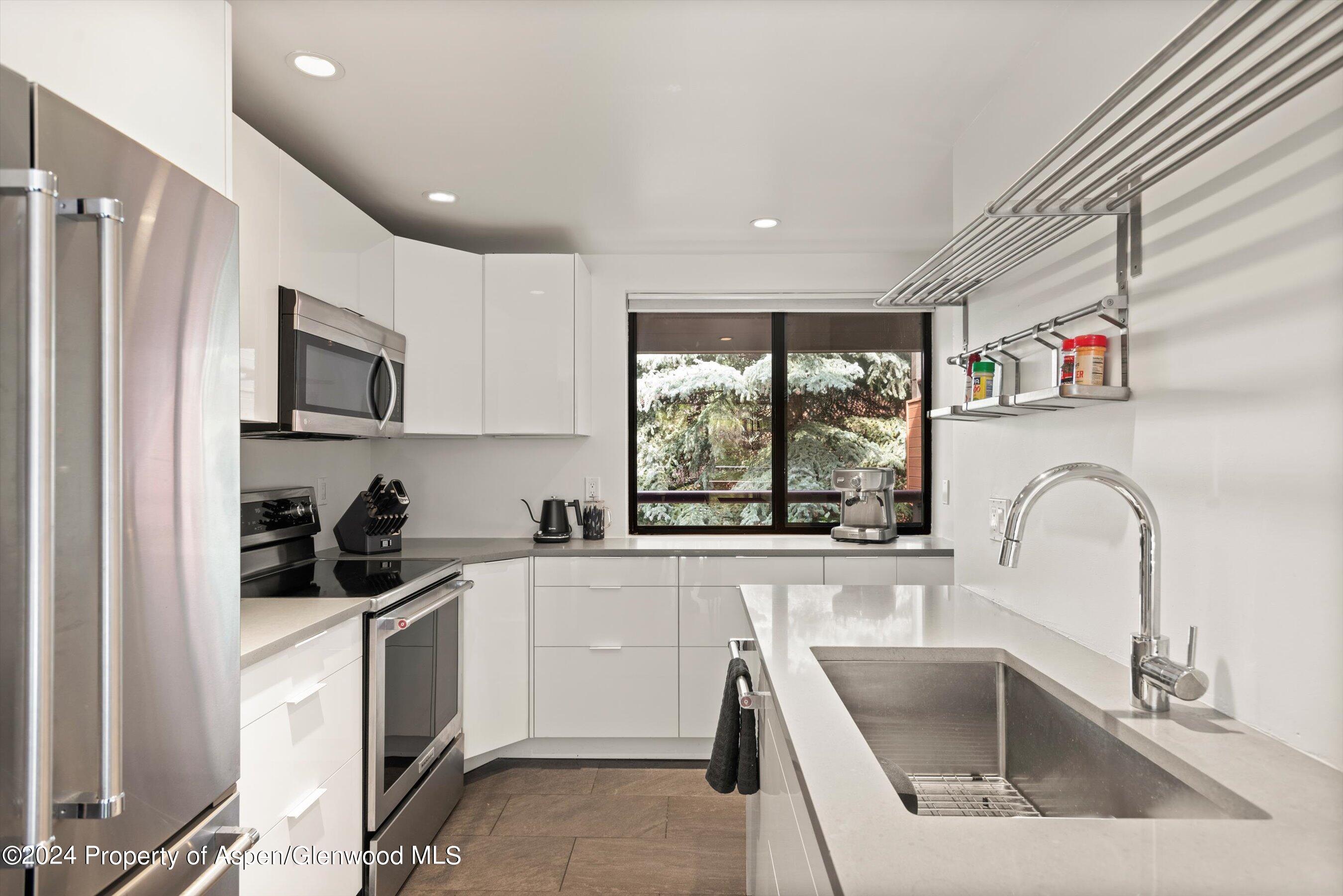 Modern white kitchen with stainless steel appliances, a large sink island, and a window overlooking evergreens.