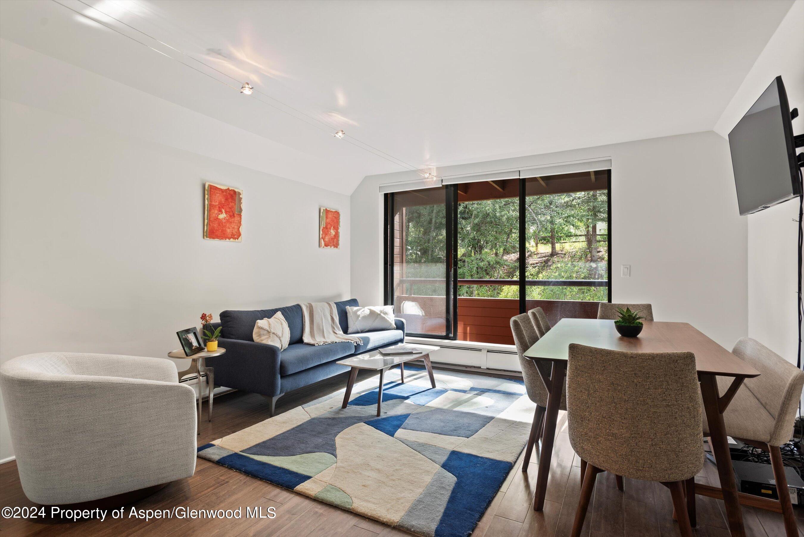 Living and dining area with a blue sofa, geometric rug, and wooded balcony view.