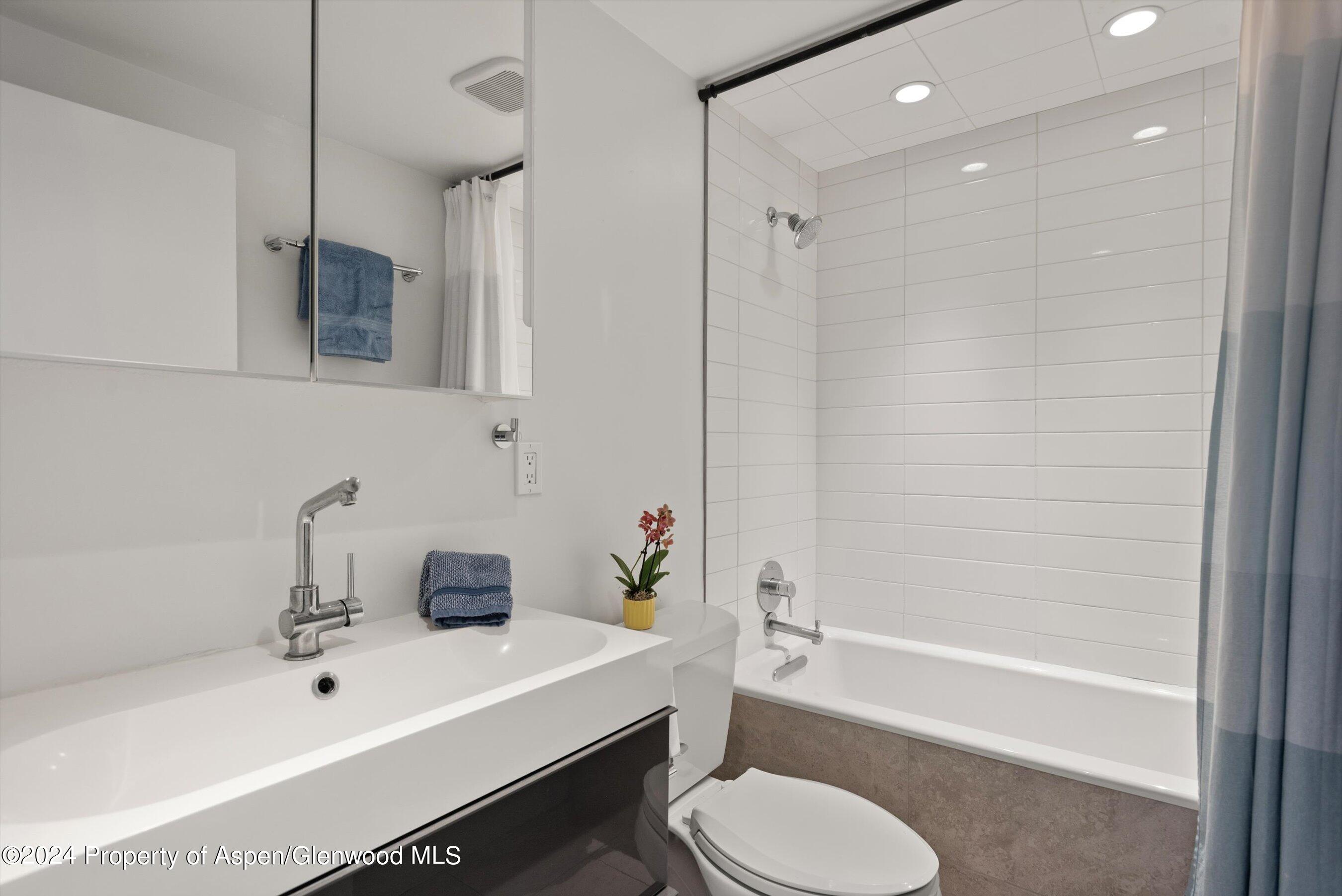 Modern bathroom with a white vanity, tiled tub and shower, toilet, blue towels, and an orchid.