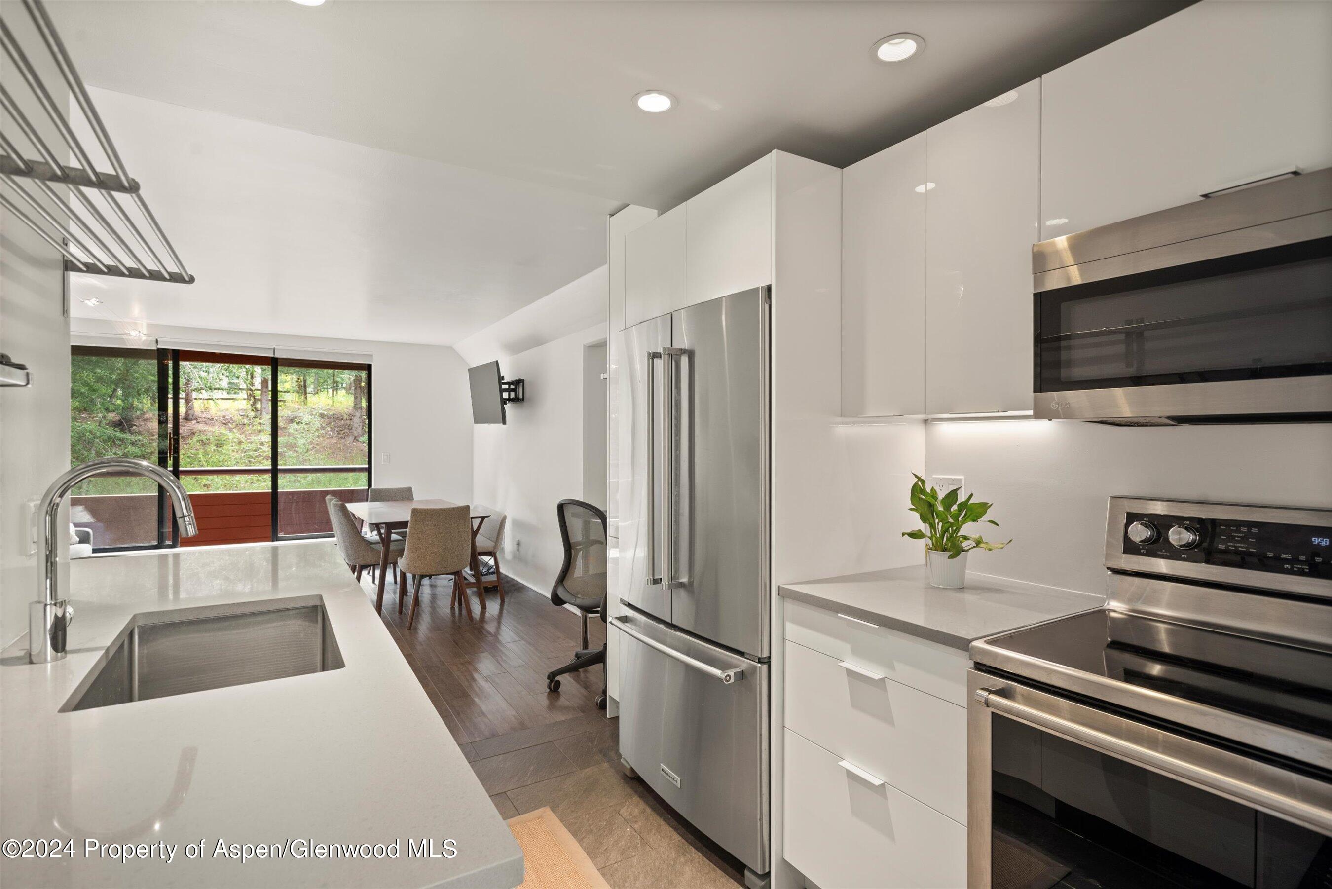 Modern white kitchen with stainless appliances, quartz counters, and dining table beside doors overlooking trees.