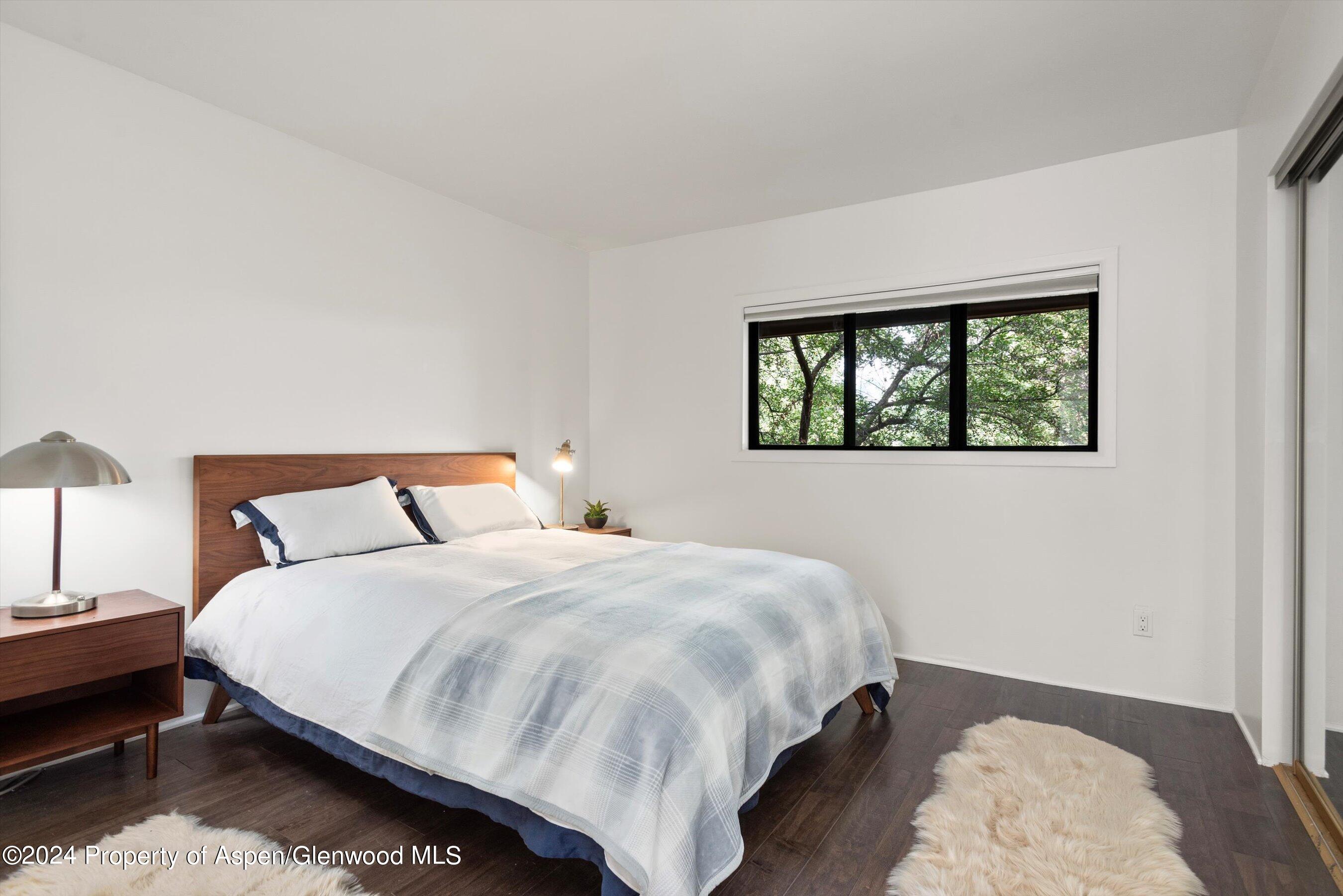Furnished bedroom with a large bed, wooden headboard, dark floors, and a window overlooking leafy trees.