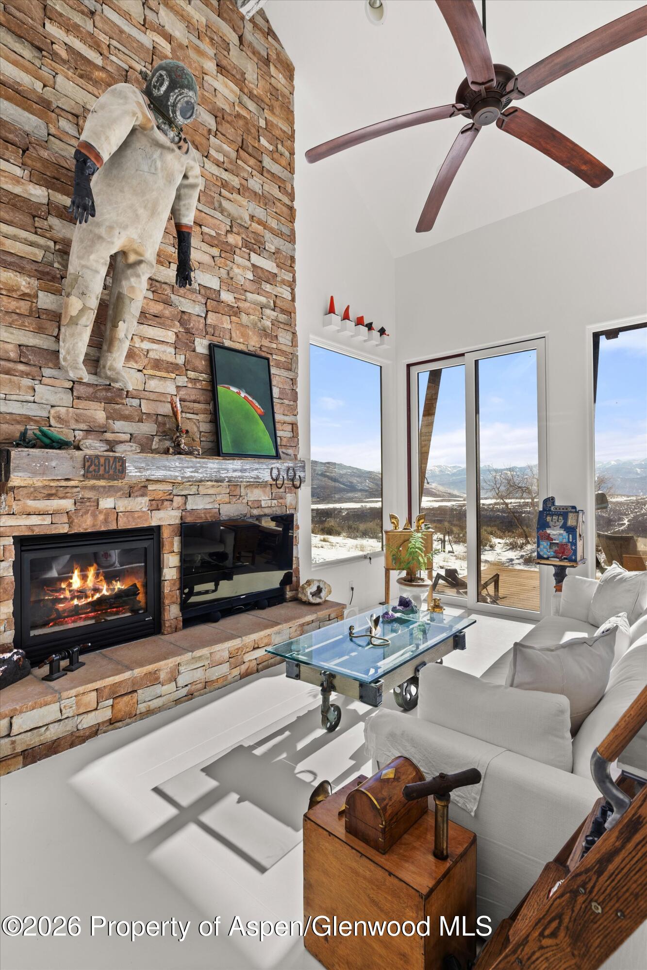 Living room with a stacked-stone fireplace, white seating, and large windows overlooking snowy mountains.