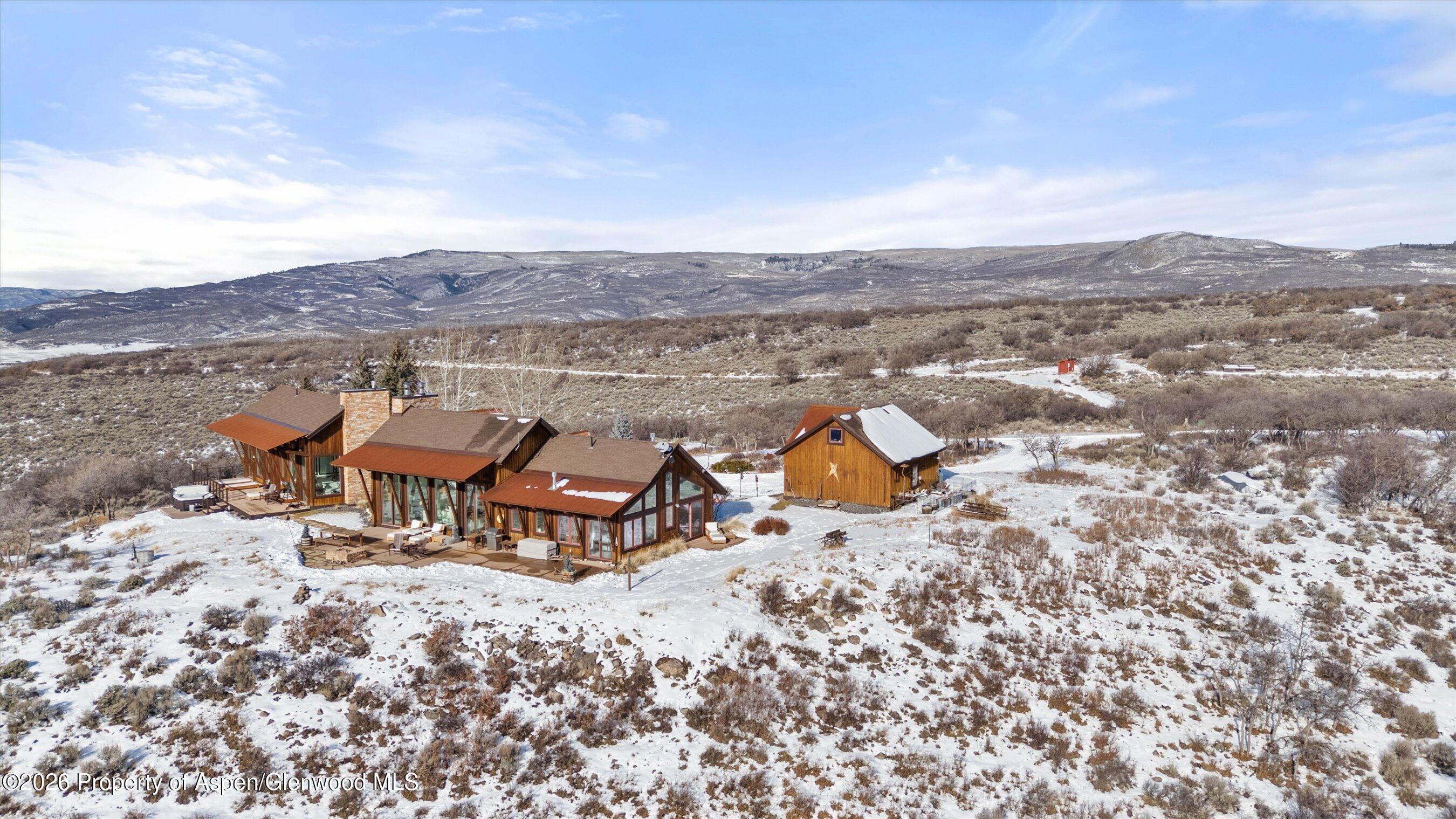 Modern timber-and-stone home with a detached outbuilding amid snow-dusted scrubland and mountain views.