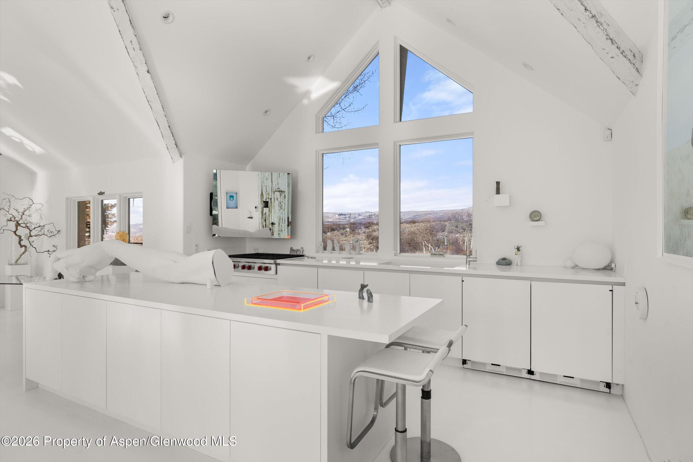 Modern white kitchen with a large island, vaulted ceiling and windows overlooking rolling hills.