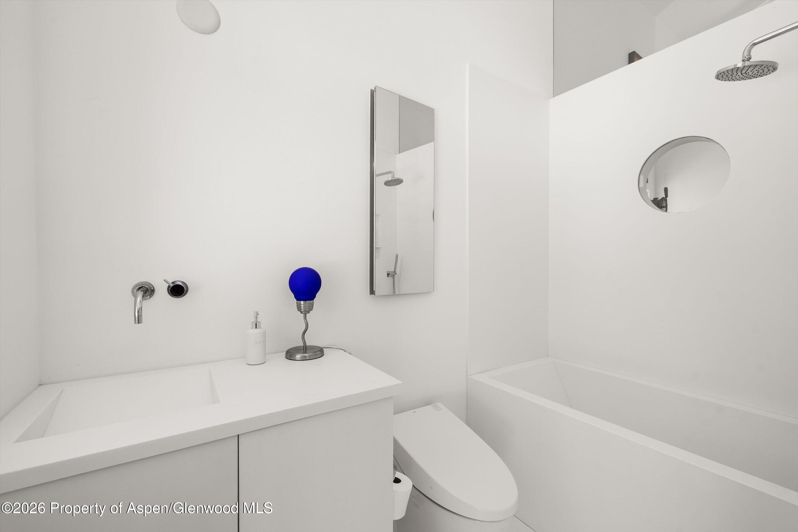 White bathroom with a built-in tub, overhead shower, integrated vanity sink, and blue lamp.