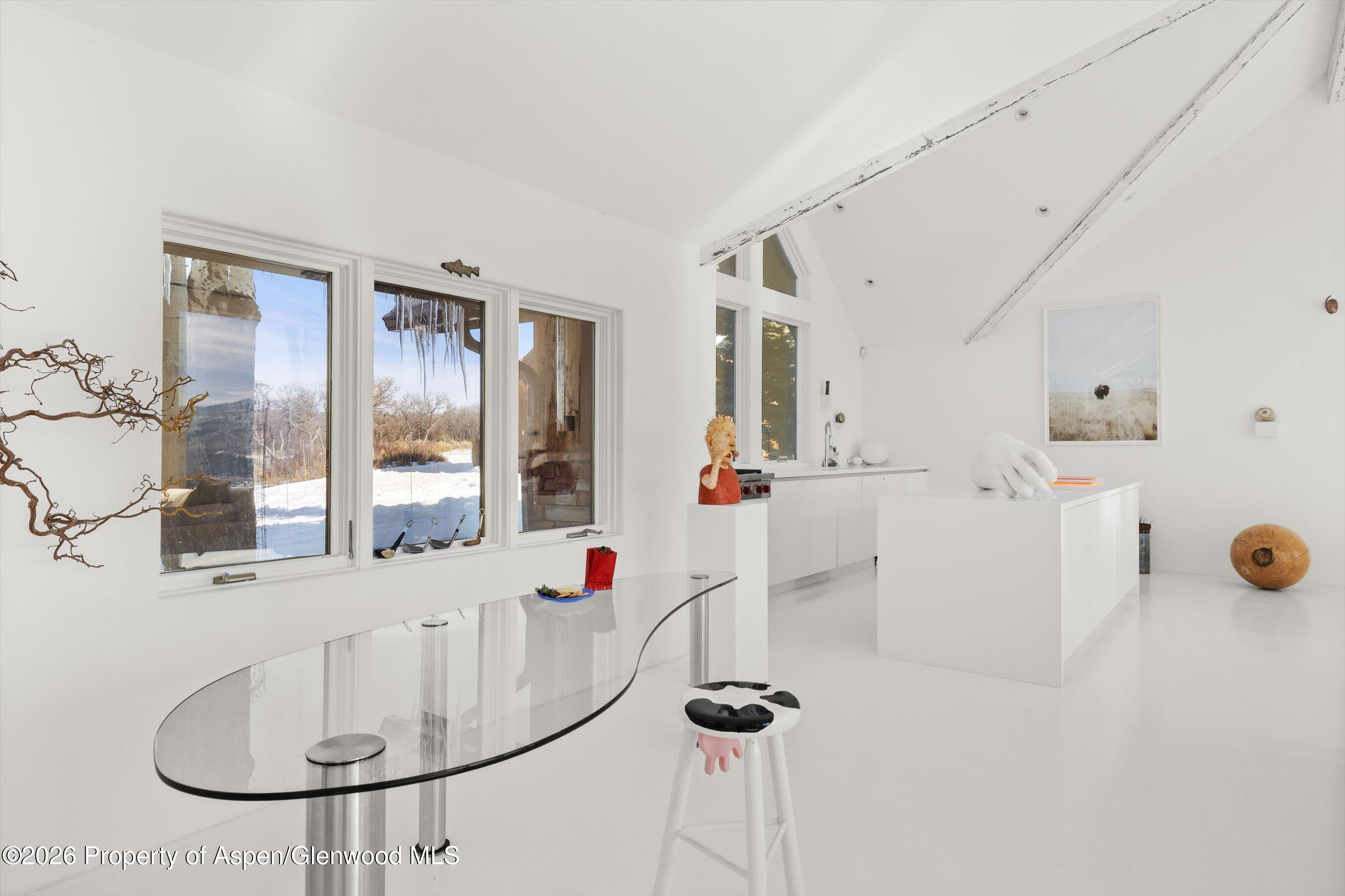 Minimalist white kitchen with vaulted ceiling, glass table, sculptural furnishings, and snowy views through the windows.