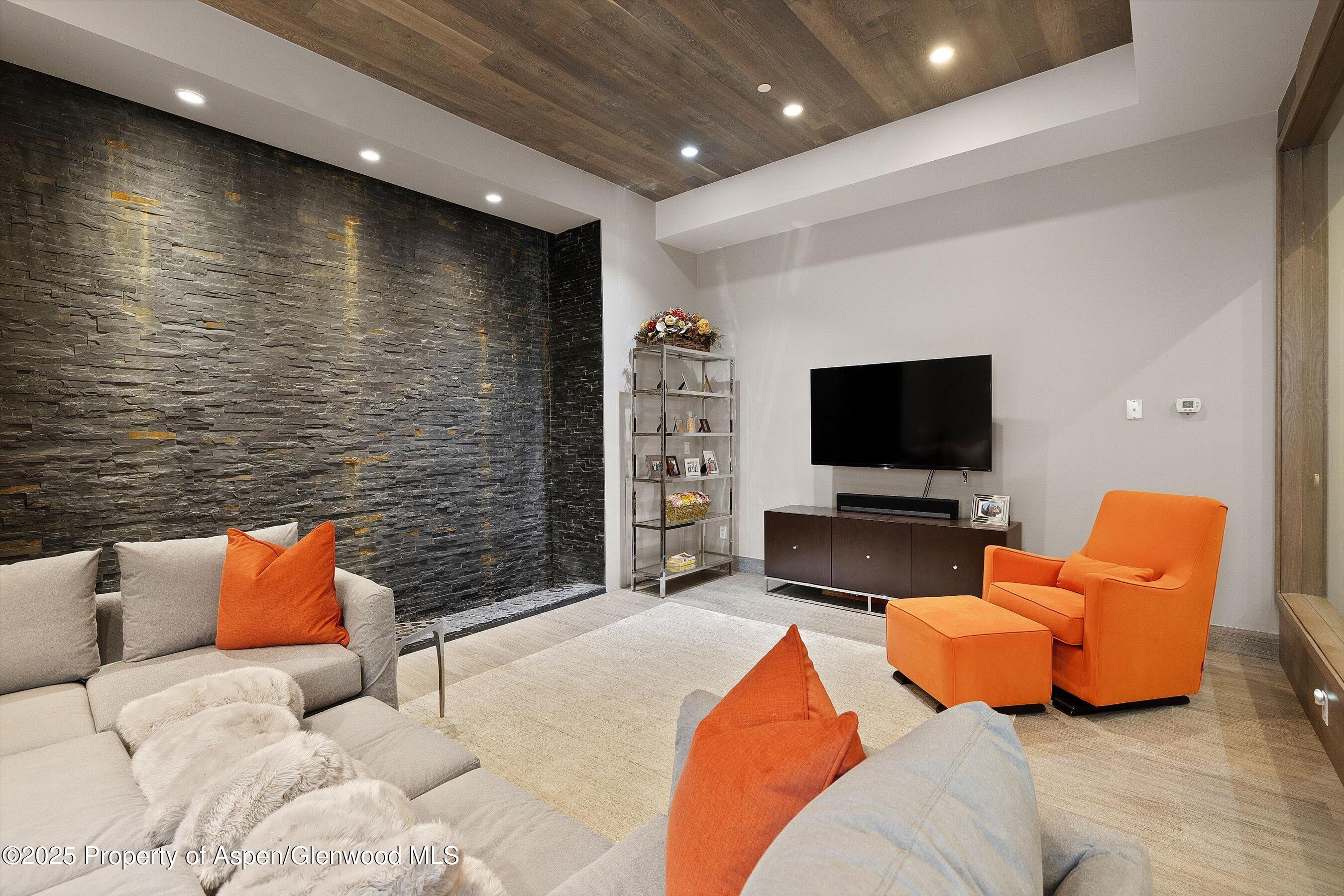 Living room with a dark stone accent wall, wood ceiling, pale sectional, and bright orange armchair.
