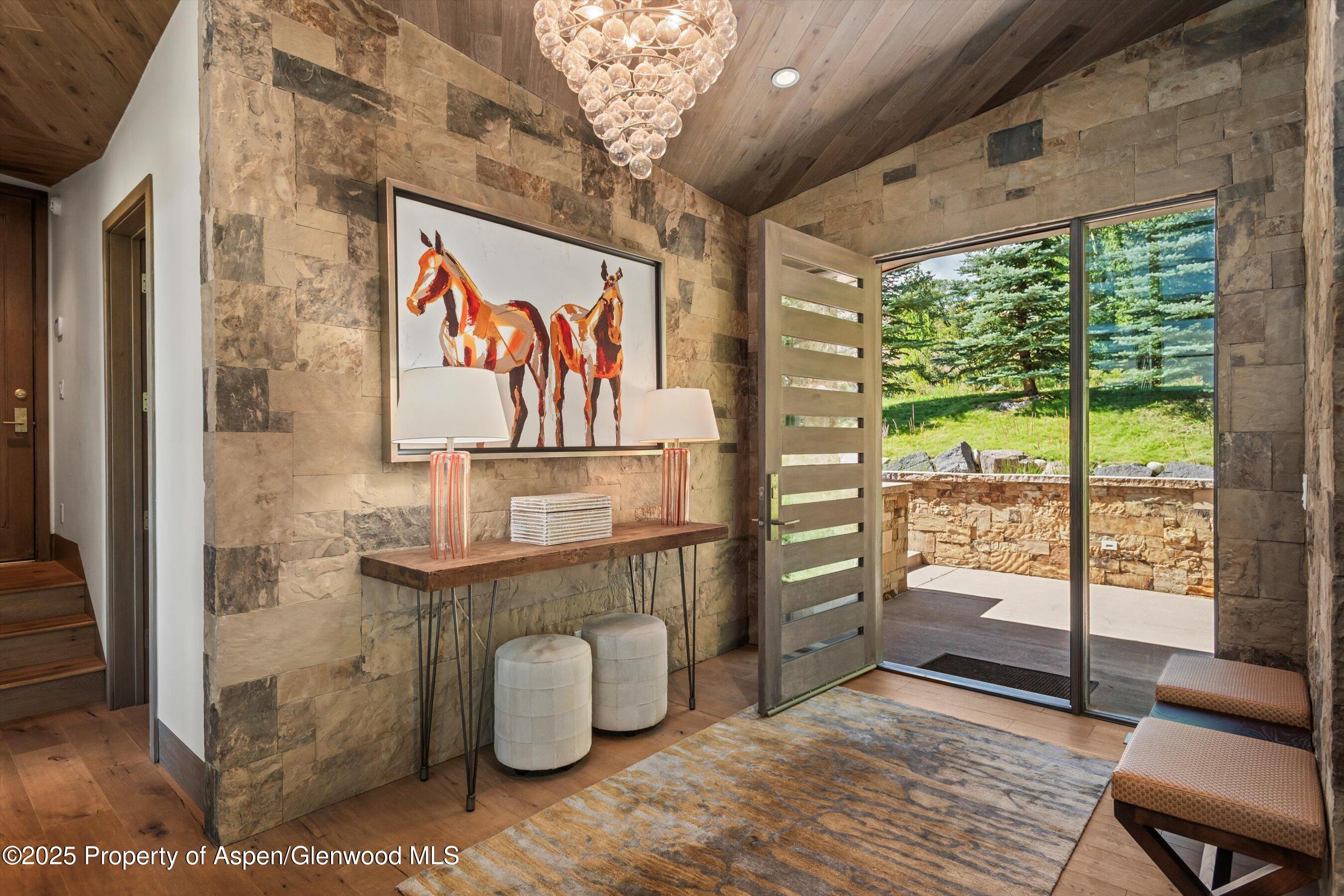 Stone-walled entryway with horse artwork, a glass chandelier, and an open door to a sunny patio.