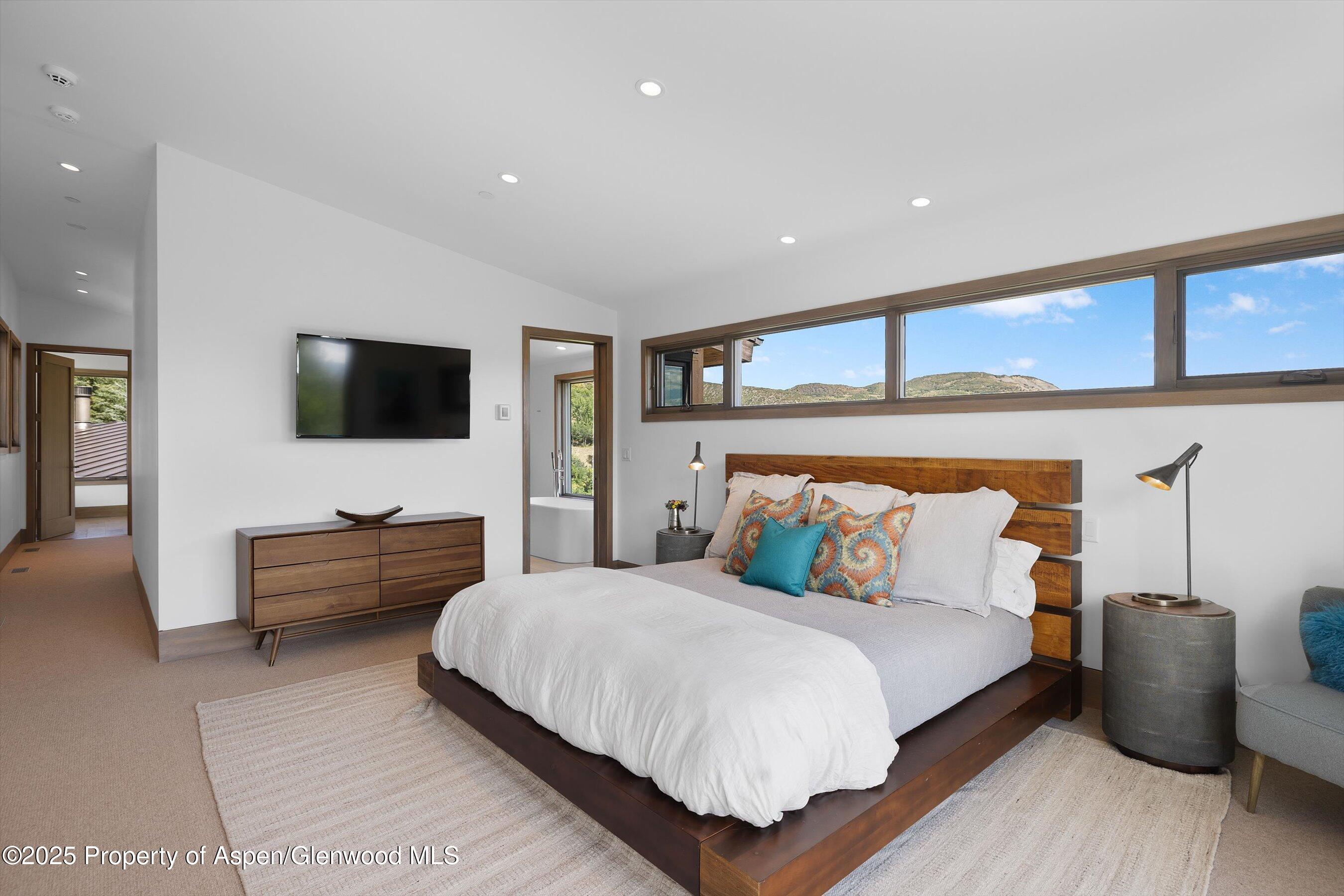 Modern bedroom with a wooden bed and long windows overlooking mountain hills.