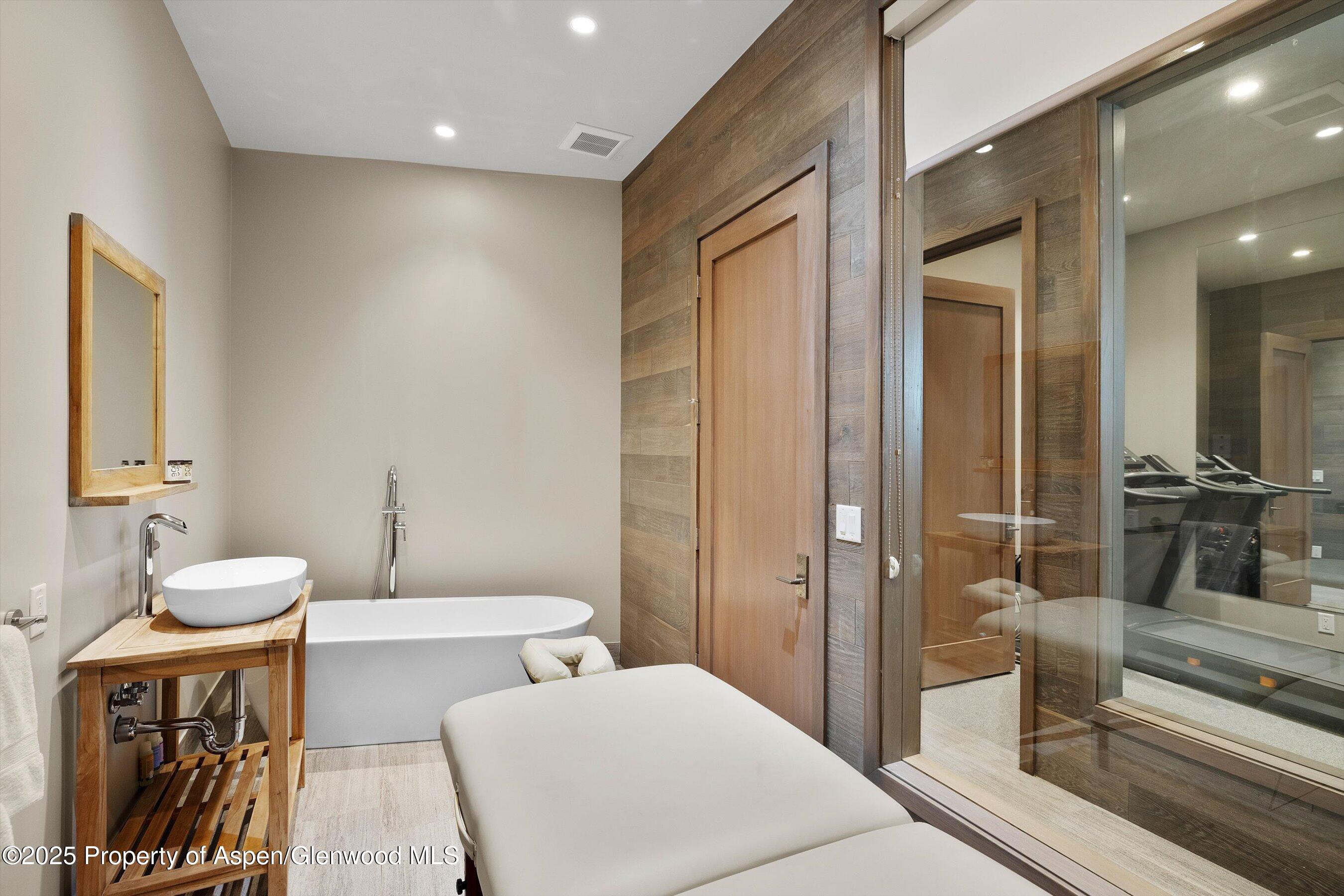 Modern bathroom with a freestanding tub, wooden vanity, and massage table beside a glass-walled gym.