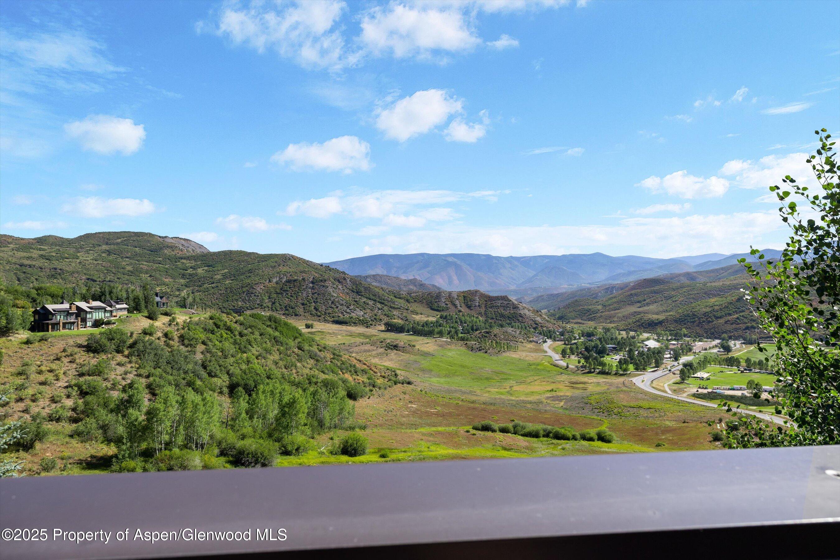 Mountain valley view with a hillside home, winding road, and deck railing in the foreground.