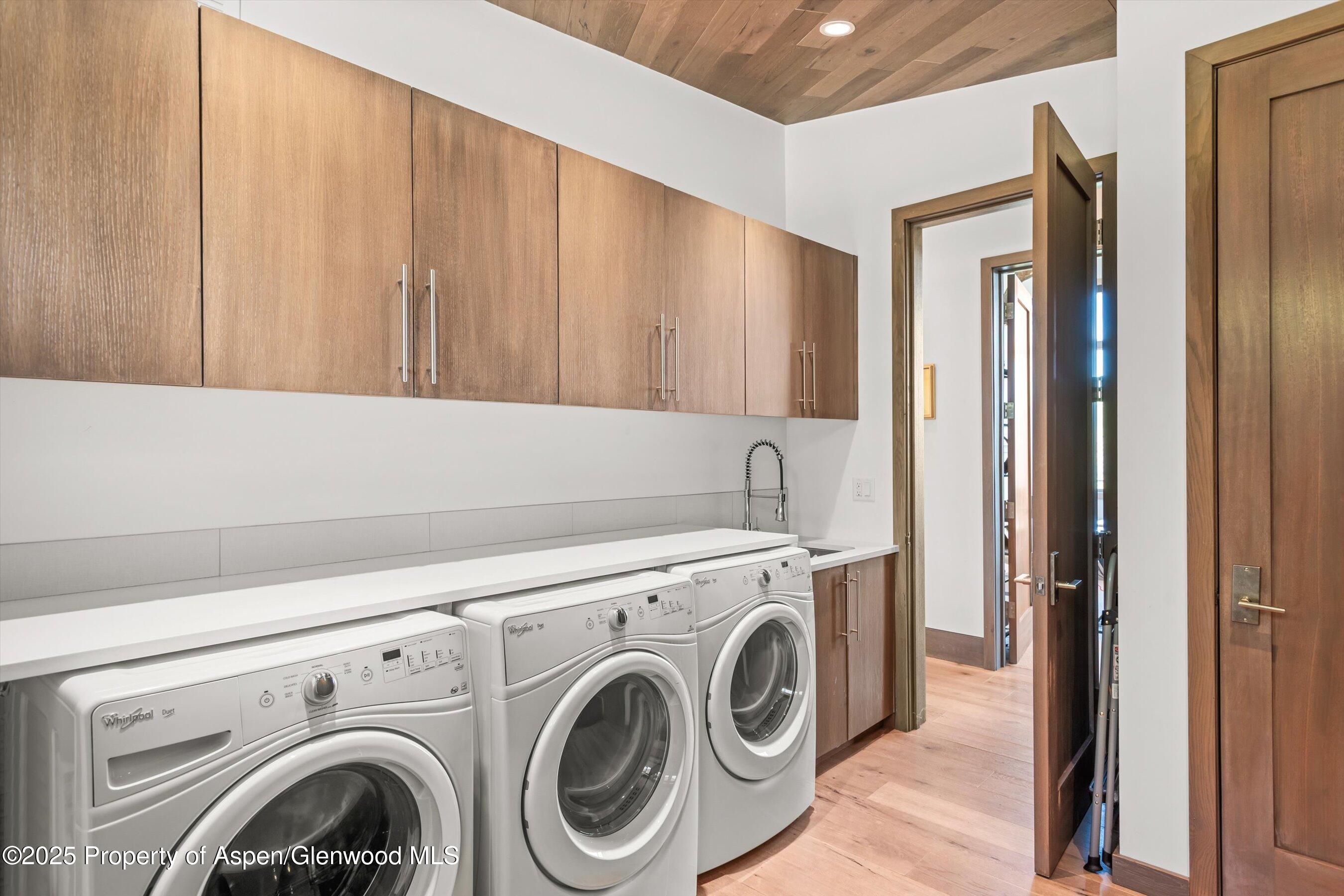 Laundry room with three front-loading machines, long countertop, and light wood cabinets.