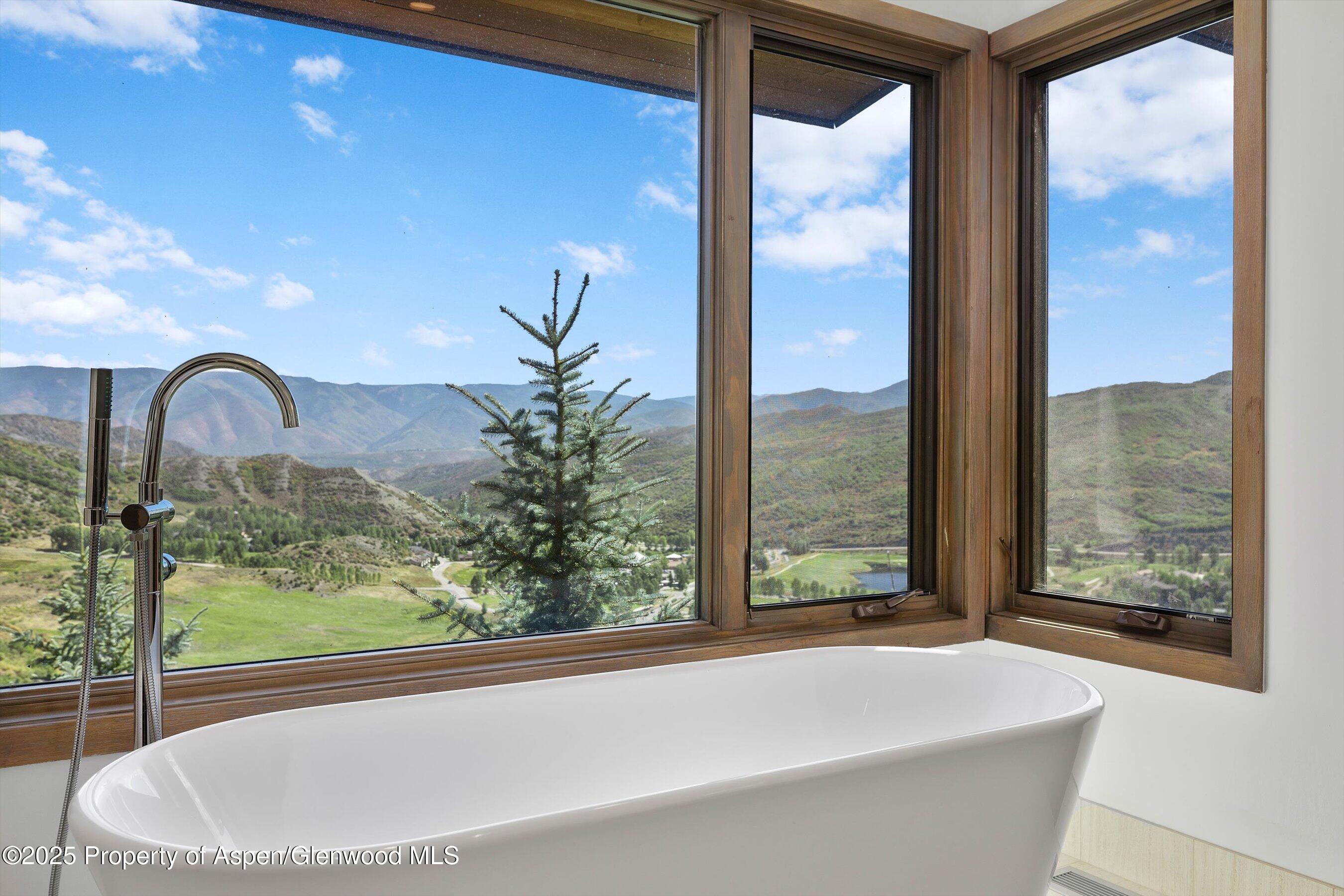 Freestanding bathtub beside broad windows overlooking a green mountain valley.