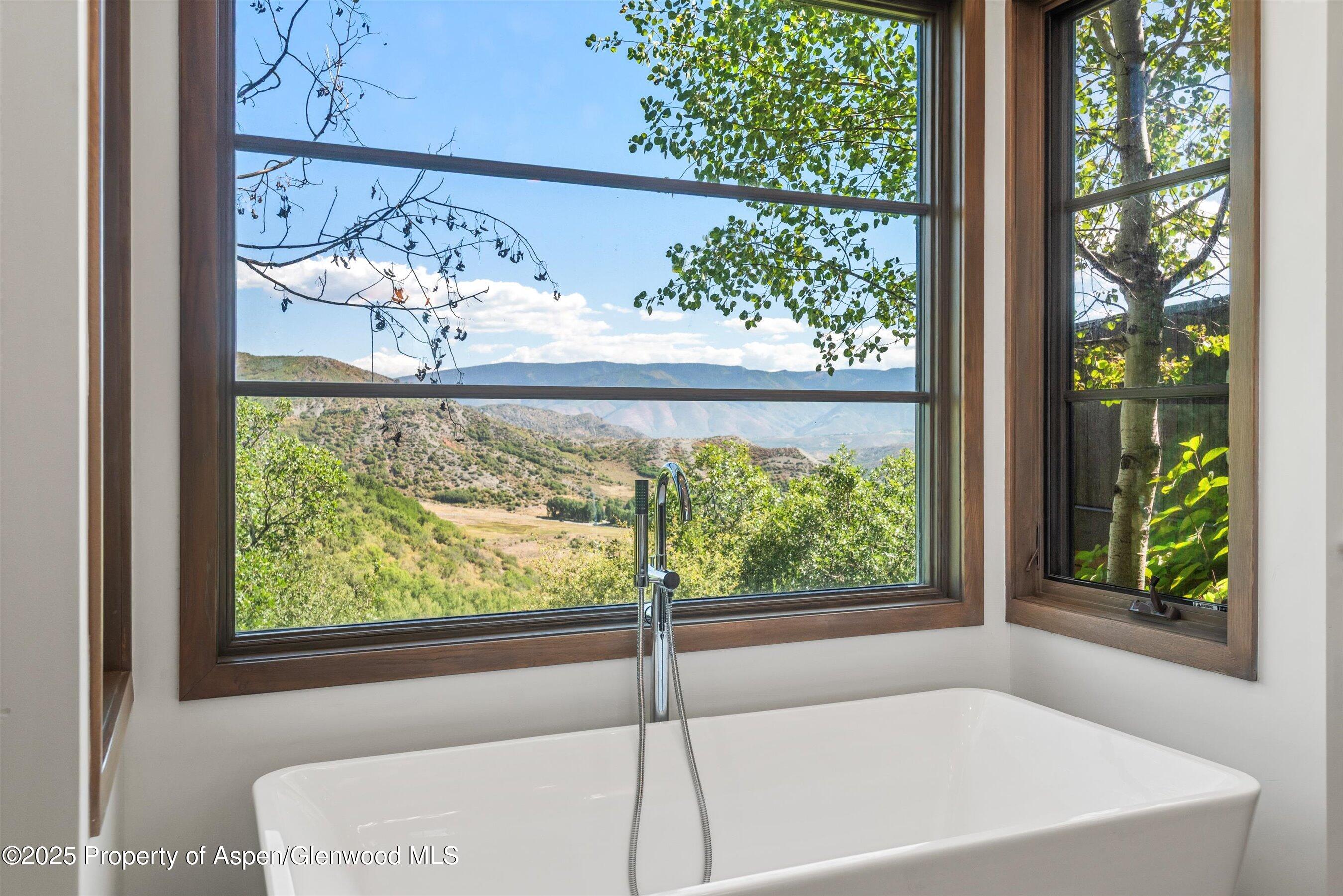 Freestanding bathtub beneath large windows with a view of green hills and distant mountains.