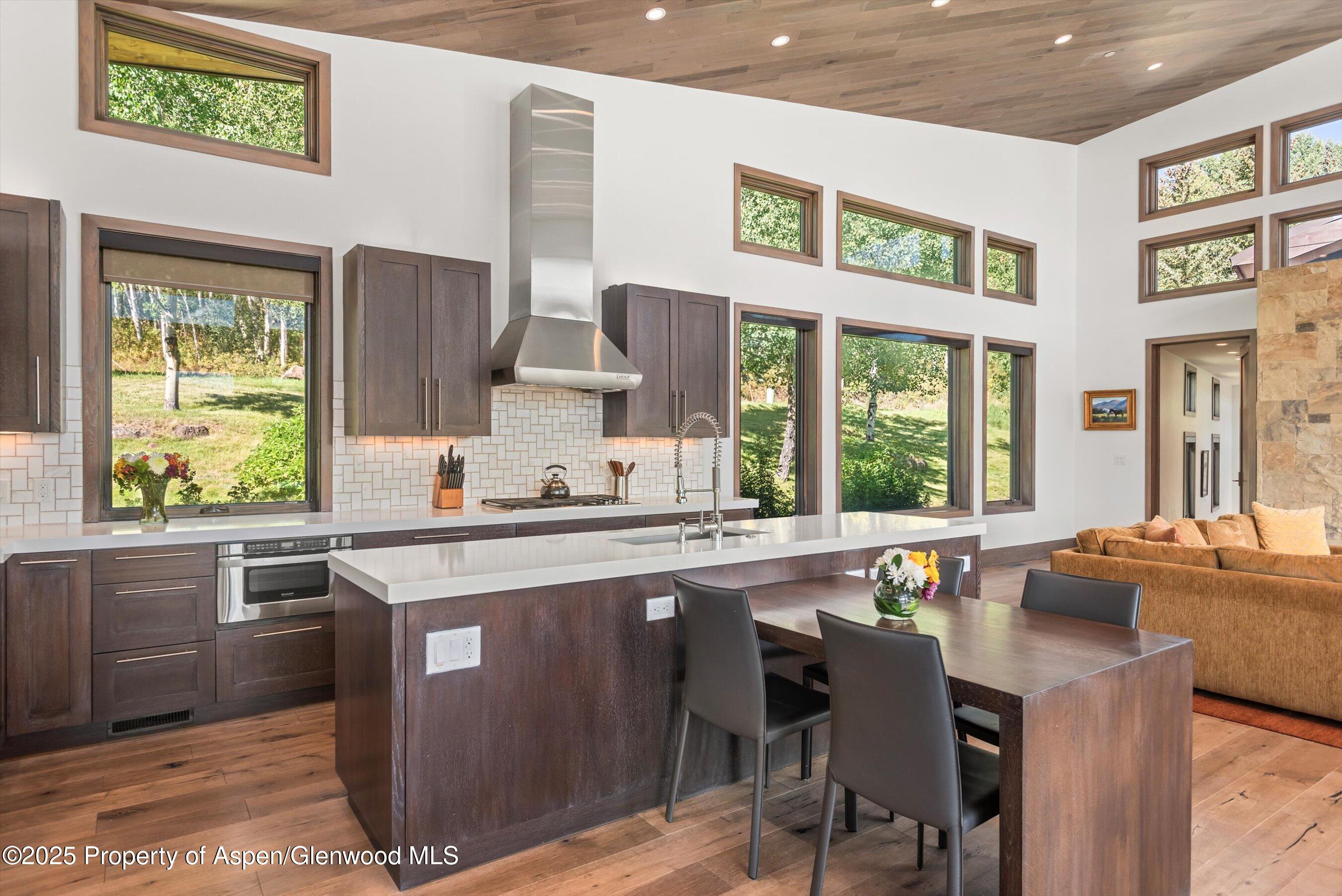 Kitchen and dining area with a large island, vaulted wood ceiling, and tall windows overlooking greenery.