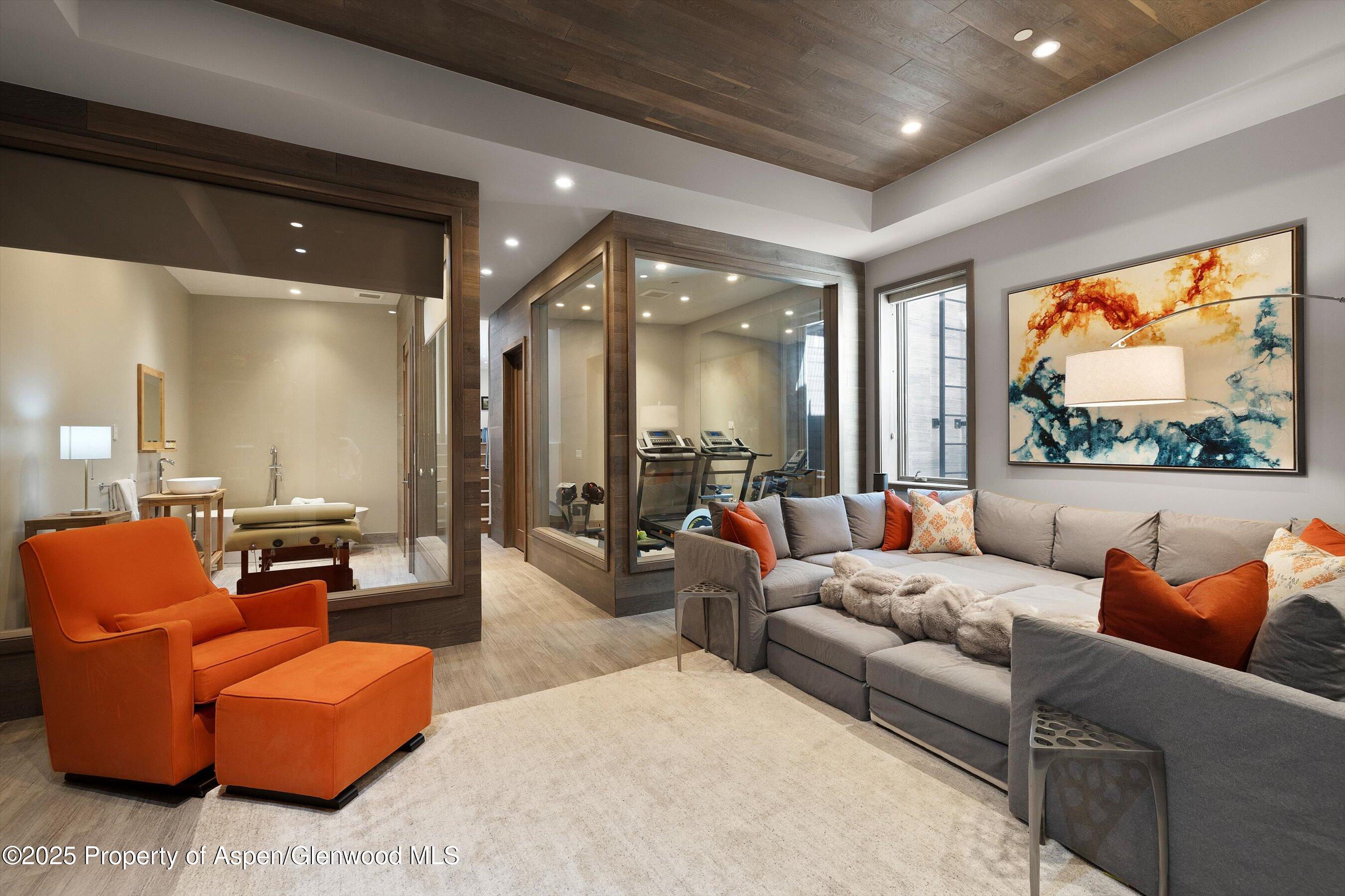 Living room with gray sectional and orange chair beside a glass-enclosed gym and massage room.